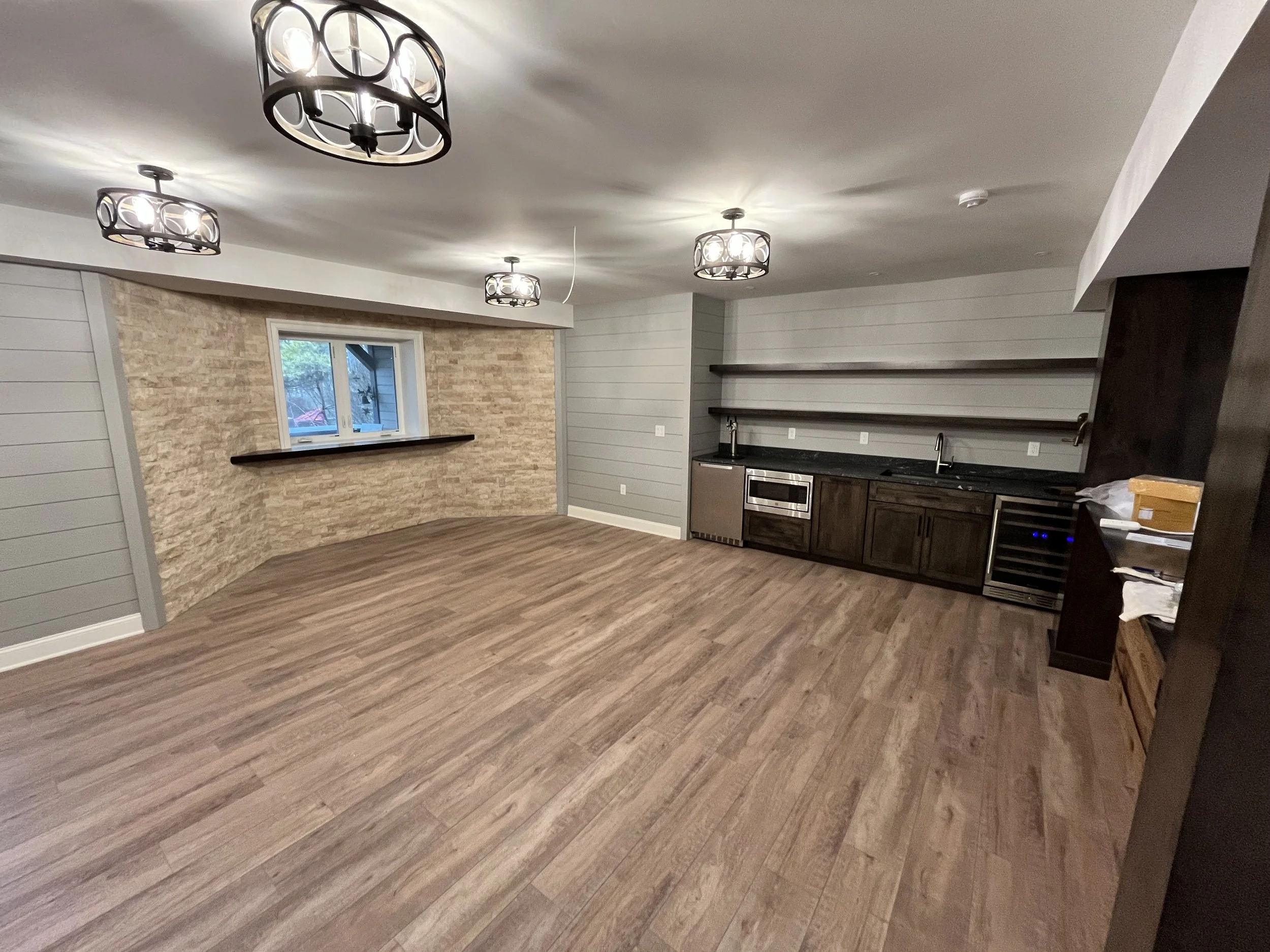A spacious modern kitchen with wood floors, gray shiplap walls, and a brick accent wall. There are four ceiling light fixtures, a small window above a countertop, and built-in appliances.