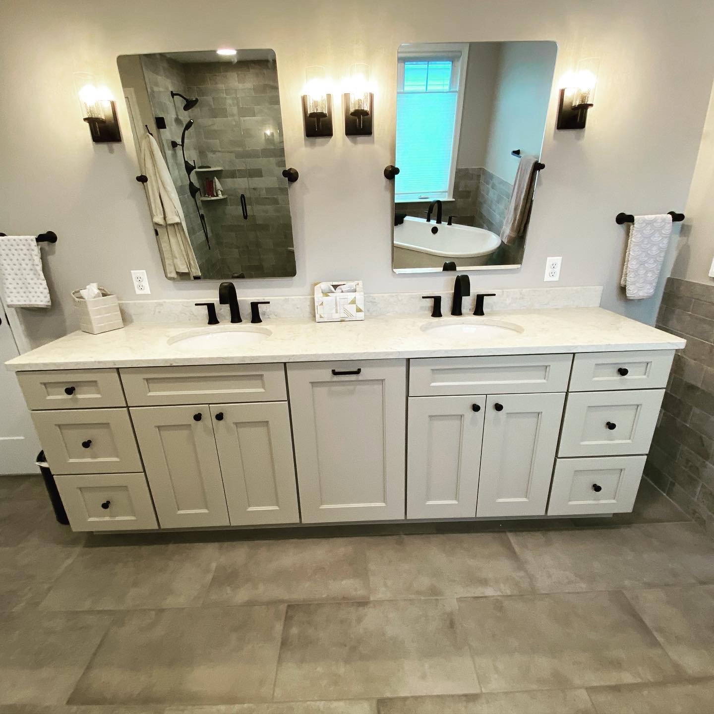 Bathroom vanity with dual sinks, black faucets, two mirrors, and wall-mounted lights. Shower visible in the reflection of the left mirror, and bathtub in reflection of the right mirror. Towel rack with towels on each side.
