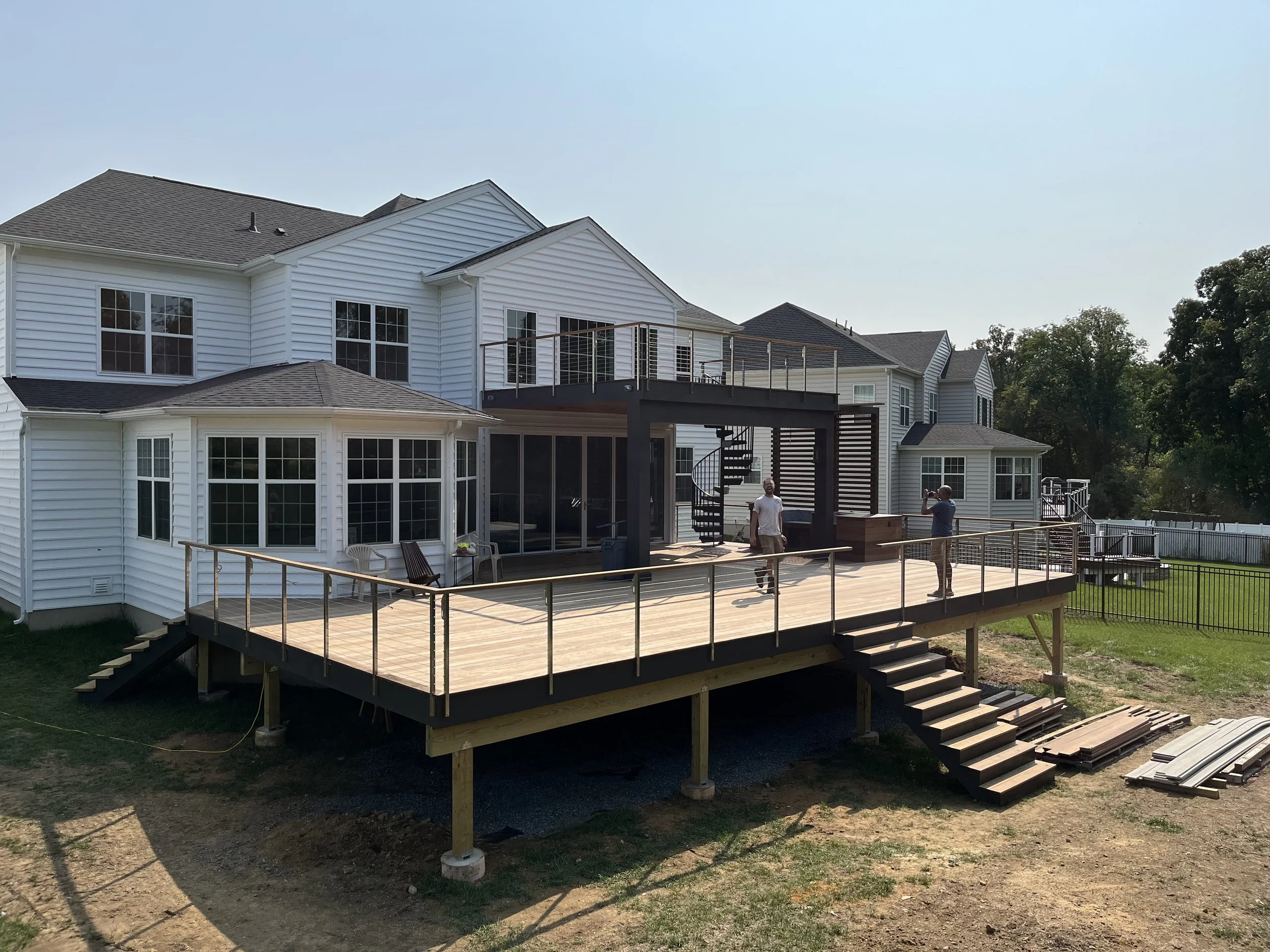 A multi-story house with white siding and numerous large windows. The house features a new wooden deck with stairs, constructed with wood and metal railings. Two men are on the deck, one standing and one taking a photo. The surrounding area includes 