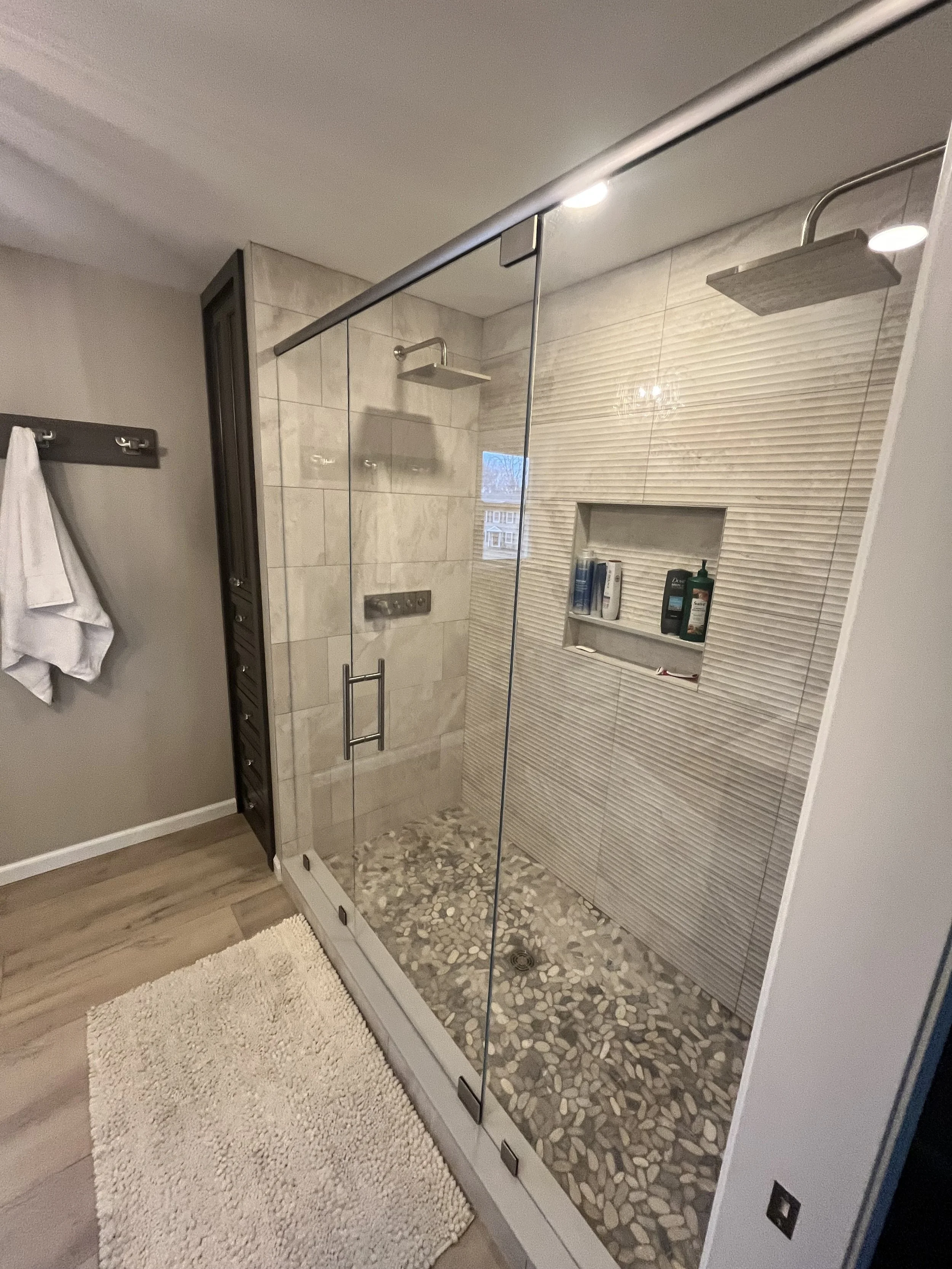 A modern bathroom with a glass-enclosed shower featuring pebble floor tiles, built-in niche with toiletries, and a dual showerhead. Towel hanging on a rack, bath mat on wood floor, and a window on the shower wall.