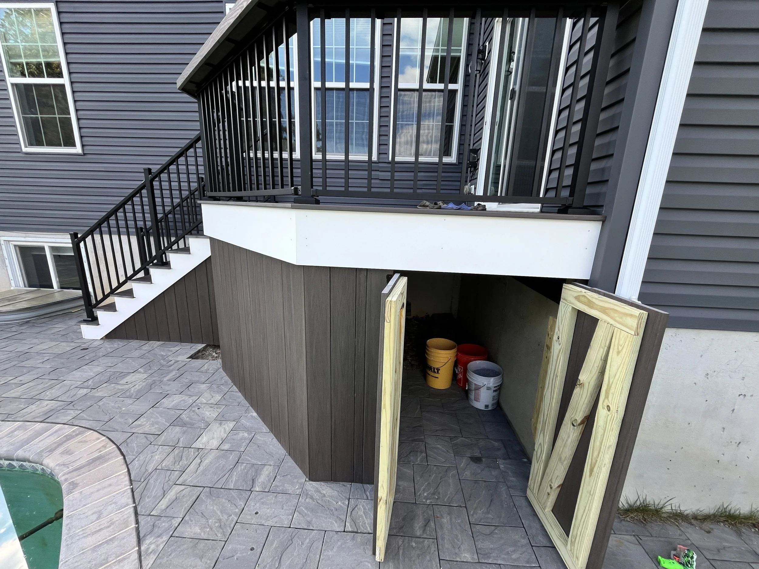Image of an unfinished outdoor balcony with a black metal railing and a small white ledge, attached to a house with dark gray siding and multiple windows. Construction supplies, including buckets and wooden planks, are stored underneath.