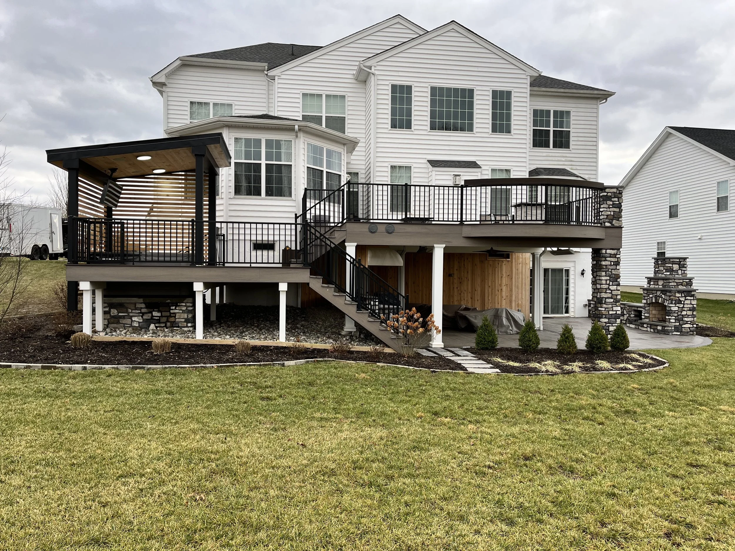 Back view of a large white house with multiple windows and a wooden deck with black railing, stairs leading down to a lawn, and a covered seating area with a wooden ceiling.