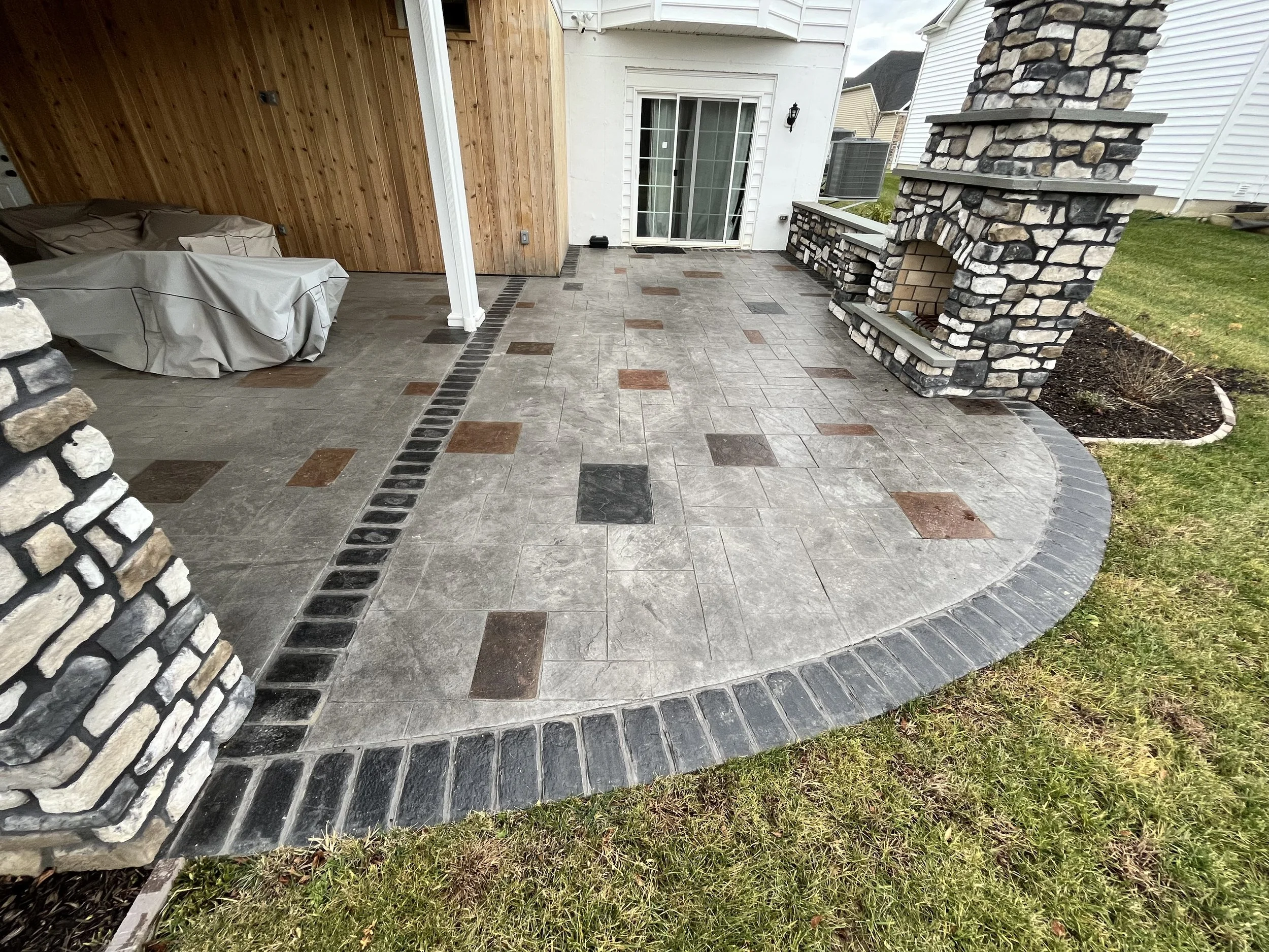 A backyard patio area with multi-colored stone tiles, a stone fireplace, a wooden wall, and a sliding glass door leading into the house. There is a covered outdoor furniture piece in the left corner.