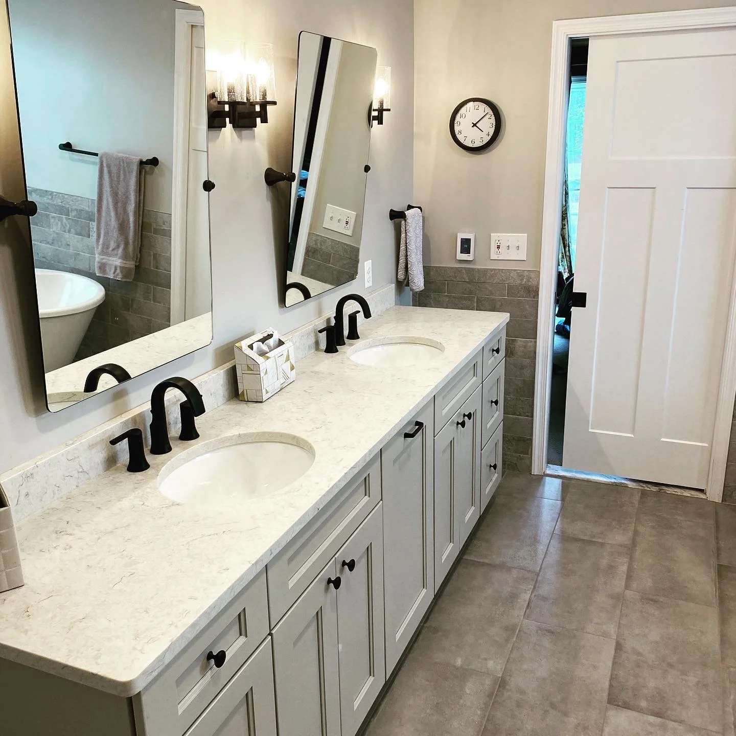 Double bathroom vanity with two oval sinks, black faucets, and a marble countertop. There are two mirrors, a wall clock, and towels hanging on black bars. The bathroom has beige tile flooring and wall decor, with an open door leading to a room with a
