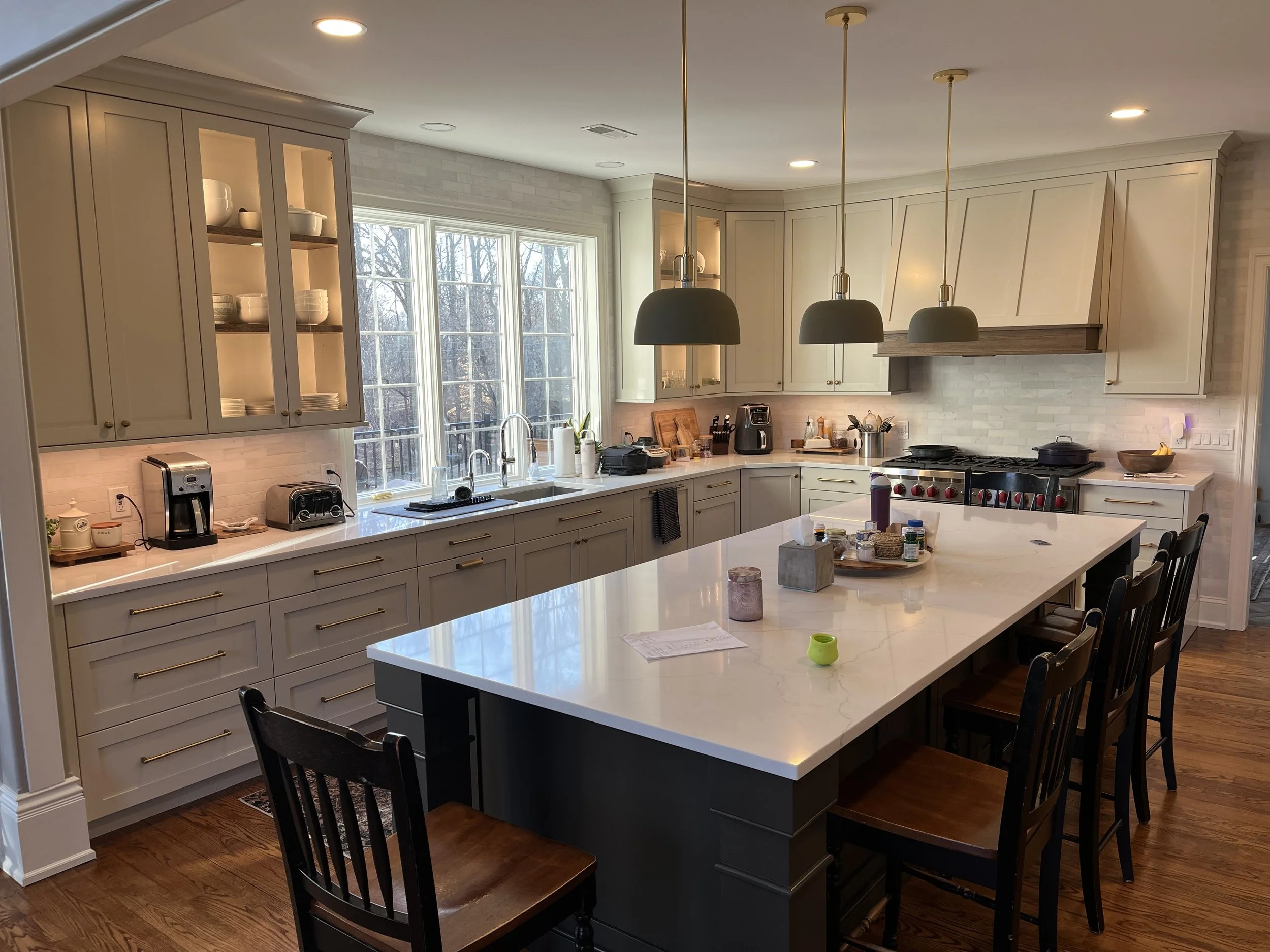 A modern kitchen with white cabinets, a large window, a white marble island, black chairs, and various appliances on the countertops.