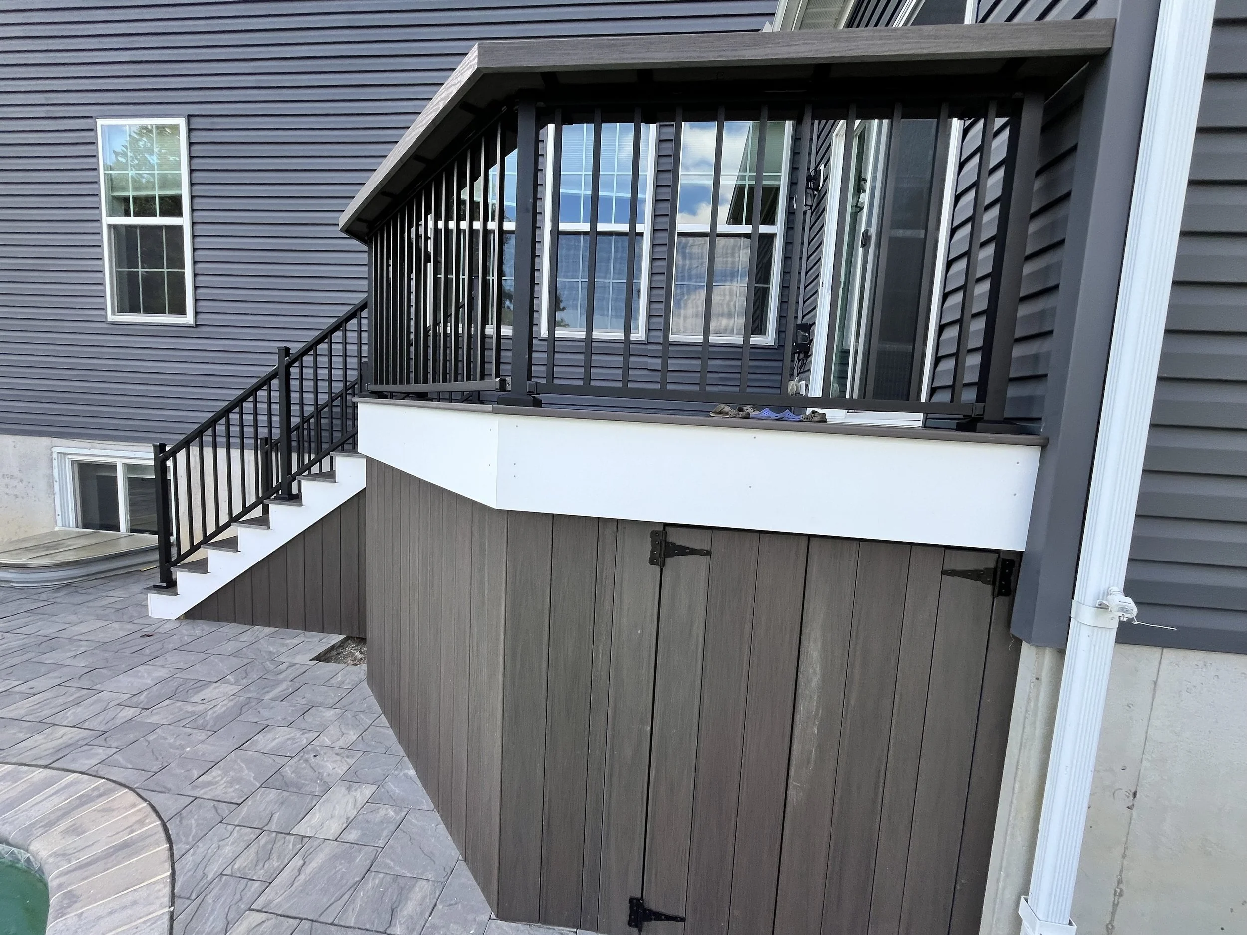 Exterior view of a house showing a small balcony with black metal railings, gray siding, a set of stairs leading up to the balcony, and a sliding glass door.