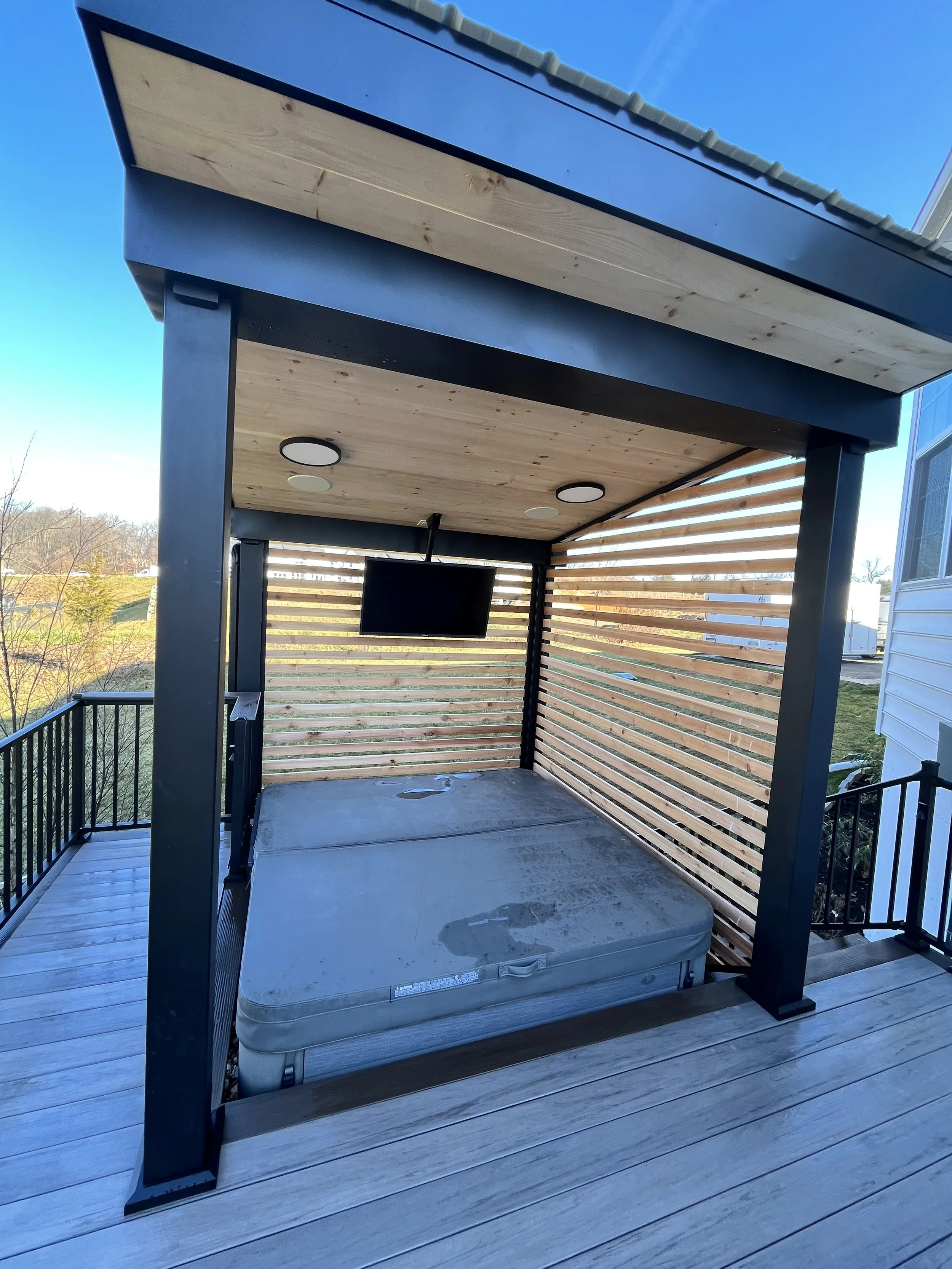 A hot tub on a wooden deck with a privacy screen made of horizontal wooden slats and a flat roof with recessed lighting. There is a remote control hanging from the roof's support beam.