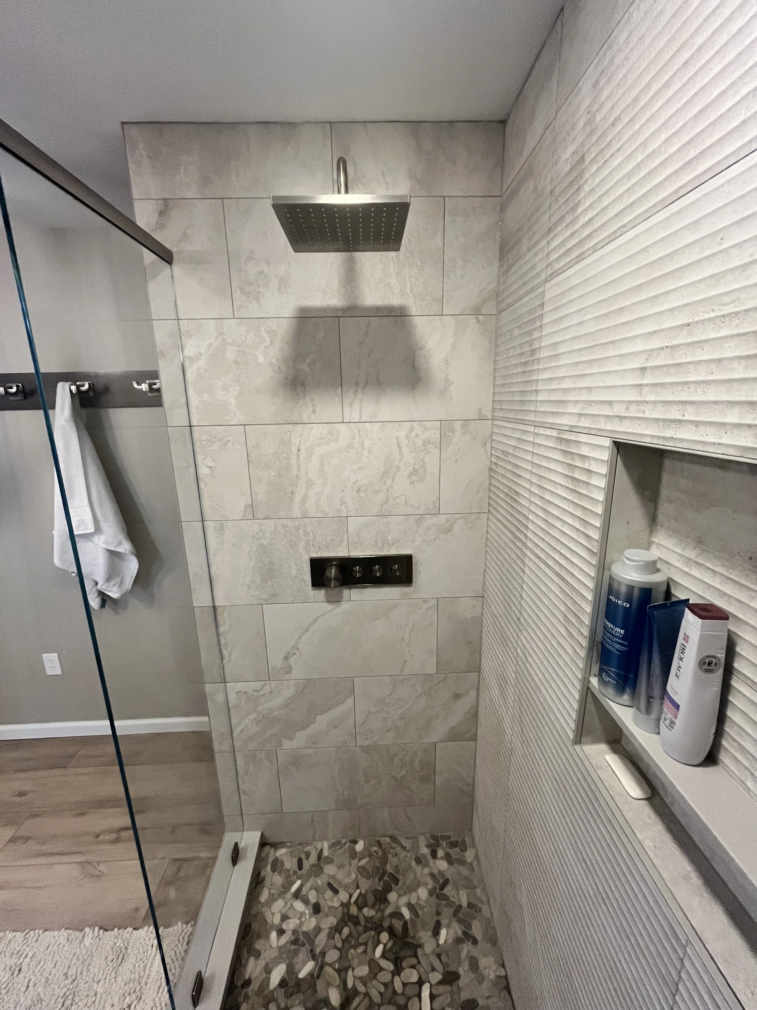 A modern bathroom with beige marble tiles on the wall, a wall-mounted showerhead, a wall-mounted control panel, and bathroom products on a white shelf.