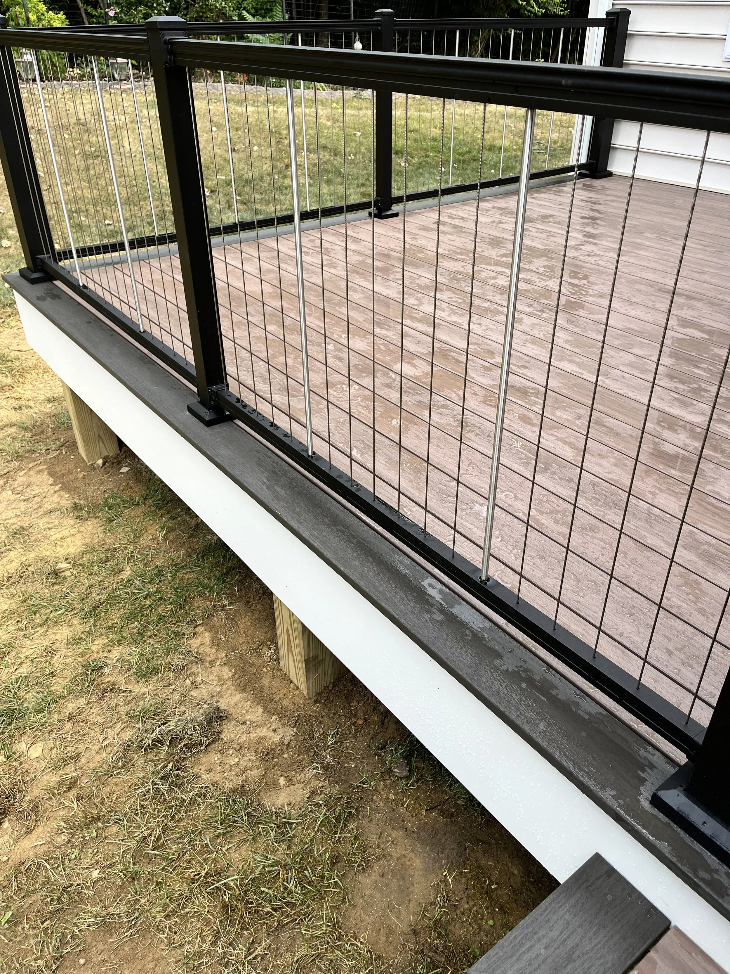 A wooden porch with black and silver metal railing, the ground below is a mix of grass and dirt.