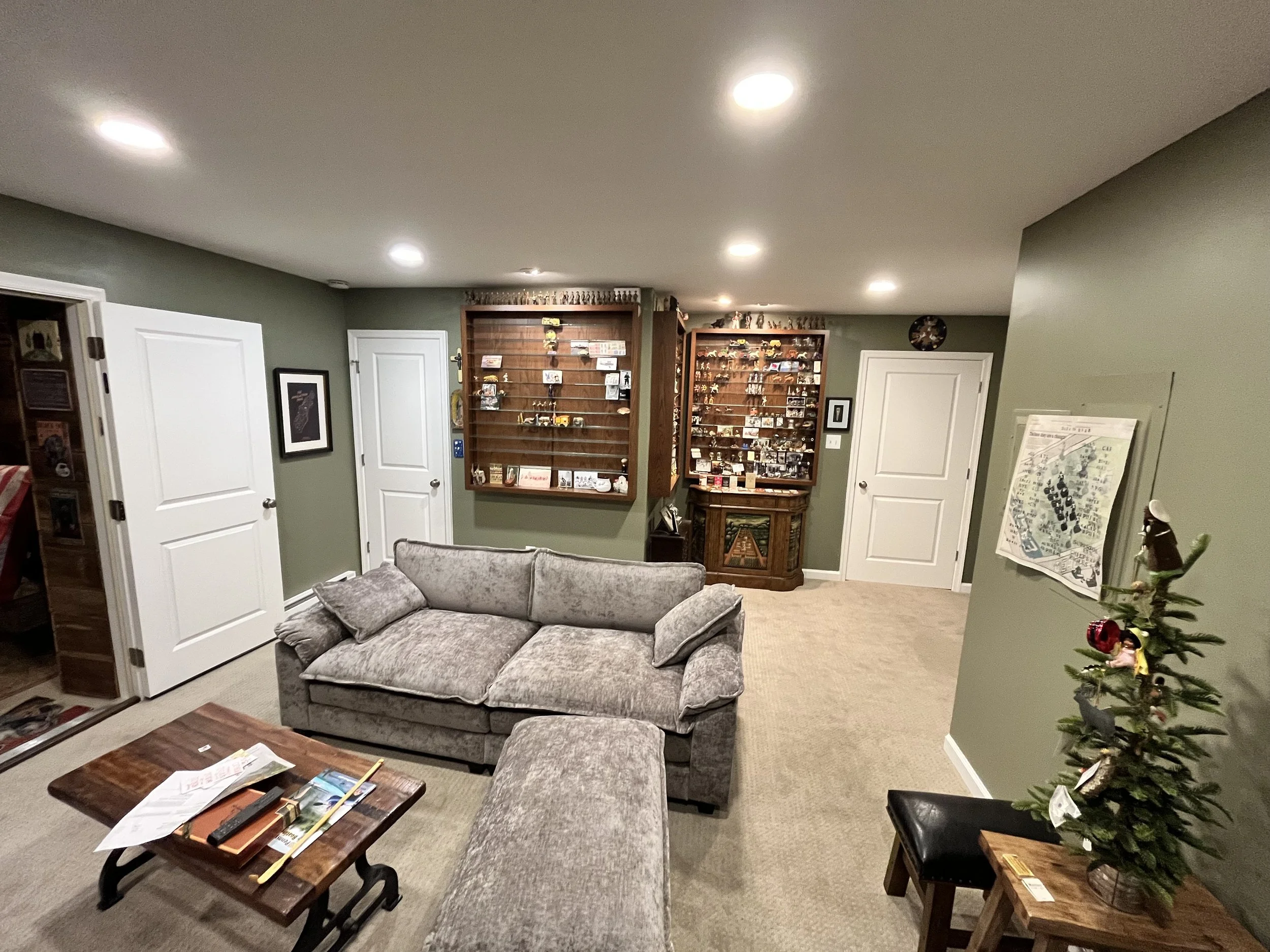 Living room with a gray sofa, a wooden coffee table with magazines, two wooden display cases filled with collectibles, and a decorated Christmas tree on the right.