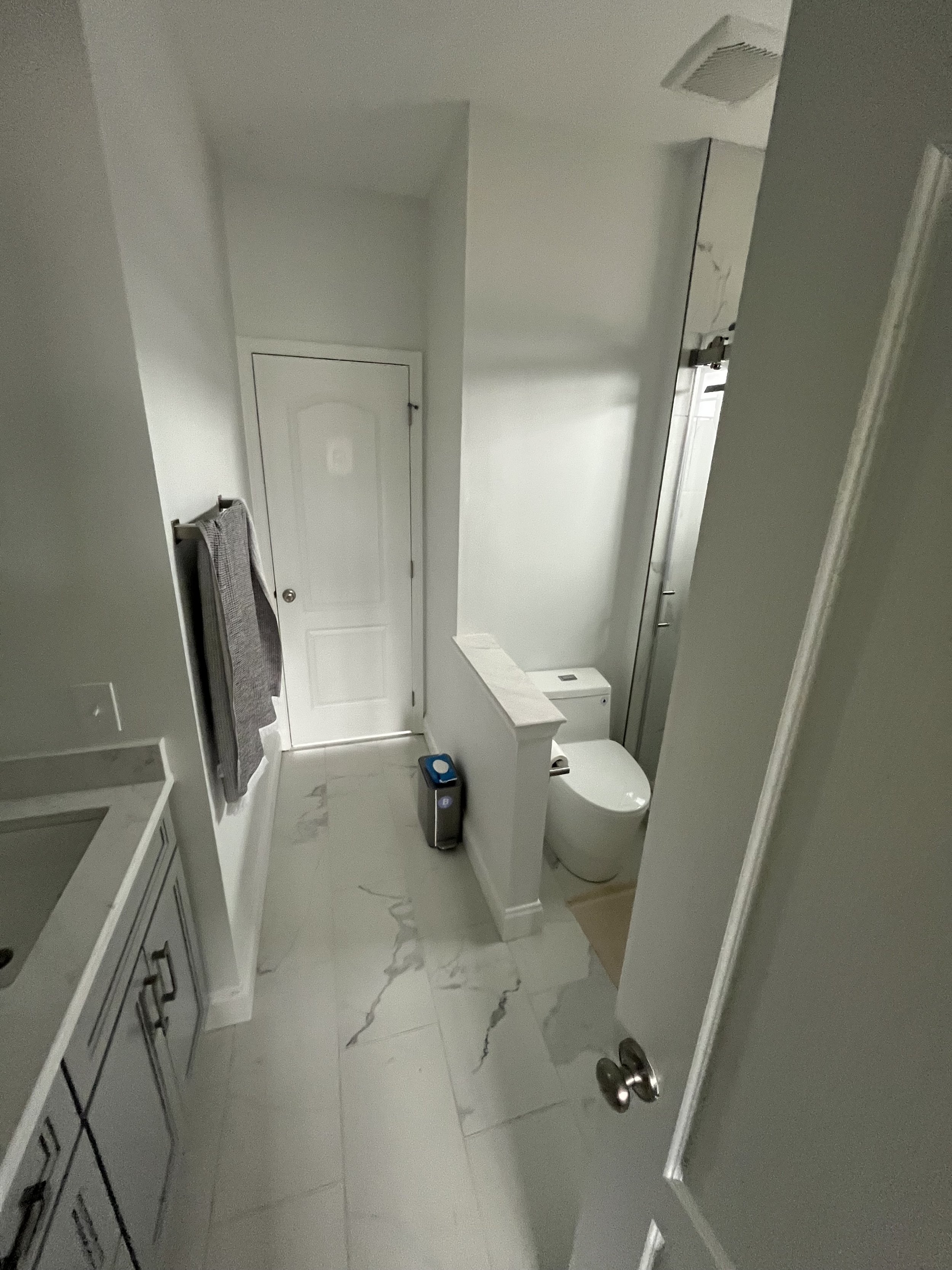 A small bathroom with white walls and floor with gray veining, a white toilet, and a white cabinet with black handles. There is a small towel hanging on a towel rack on the left wall, and a vent on the ceiling near the door.