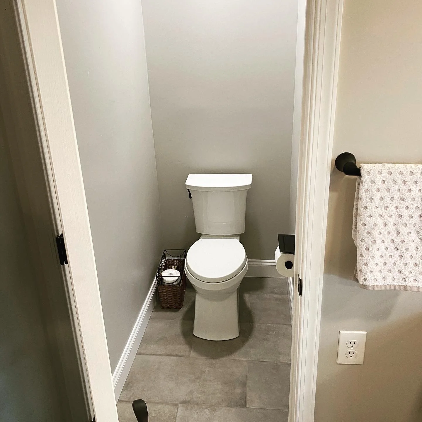 A small bathroom with a white toilet, a basket with toilet paper, and a towel hanging on a black towel rack on the right side. A black toilet paper holder is attached to the wall, and a white outlet is near the floor.