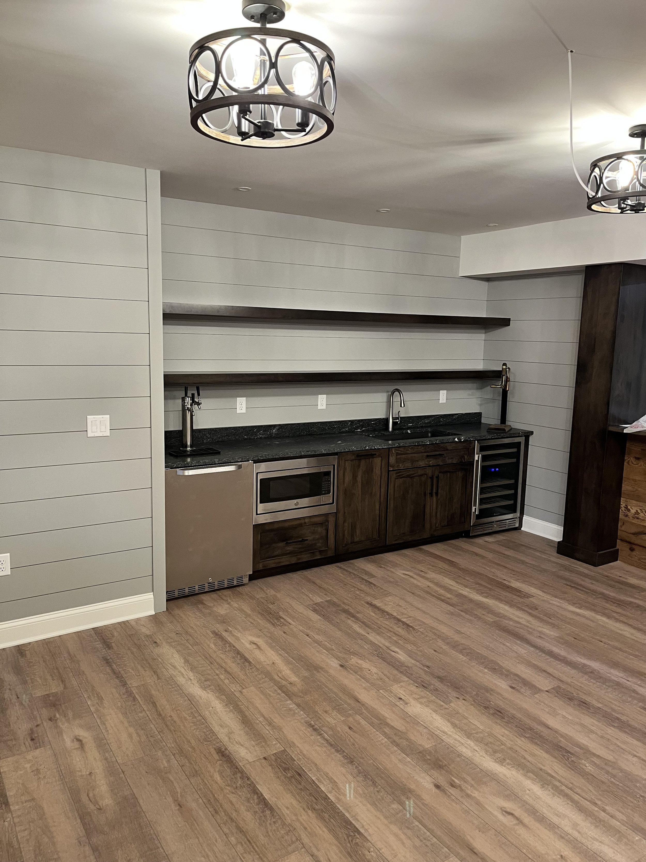 Modern kitchen with wooden cabinets, black countertop, stainless steel appliances including a microwave and wine cooler, white shiplap walls, floating shelves, and wood flooring. Two ceiling light fixtures are visible.