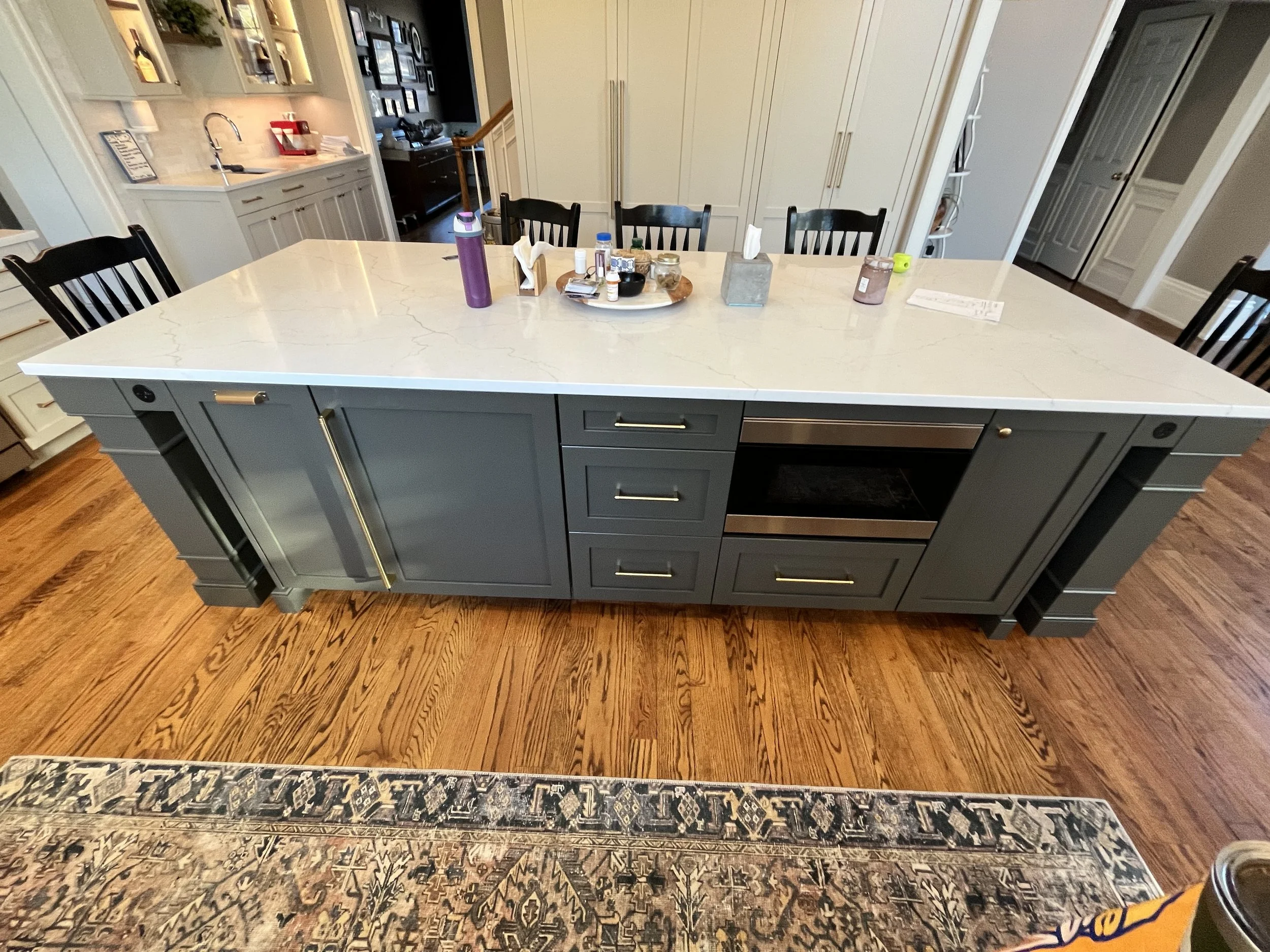 A dining area with a large marble-top table, six black chairs, a hardwood floor, and a woven area rug. The table has various items including a purple water bottle, tissue box, jars, and small containers. The background features white cabinetry, a sma