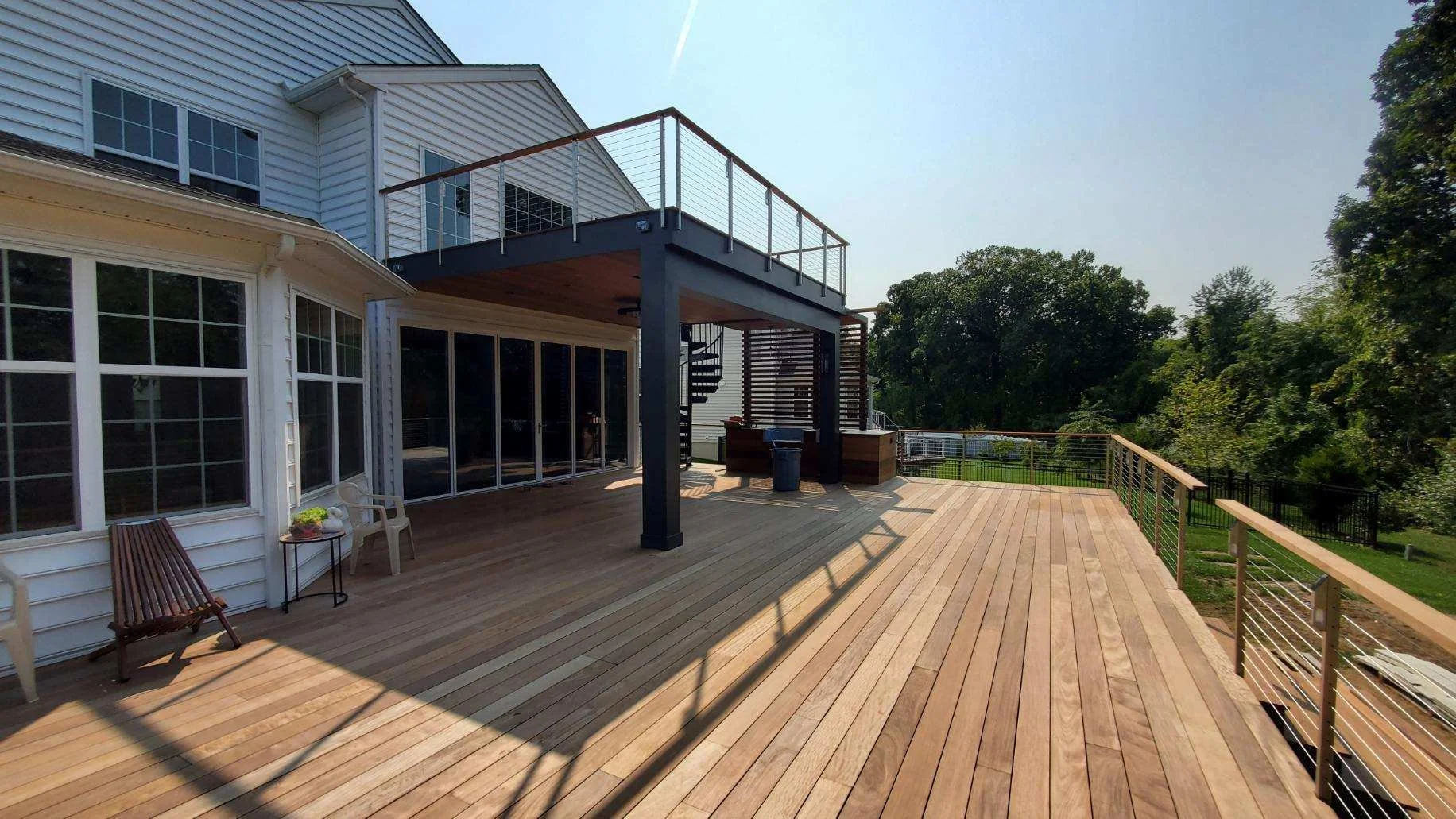 A spacious wooden deck attached to a white house with large windows and sliding glass doors. The deck has a railing, an outdoor staircase, and is partly shaded by the upper level of the house. There are trees and greenery in the background, with sunn