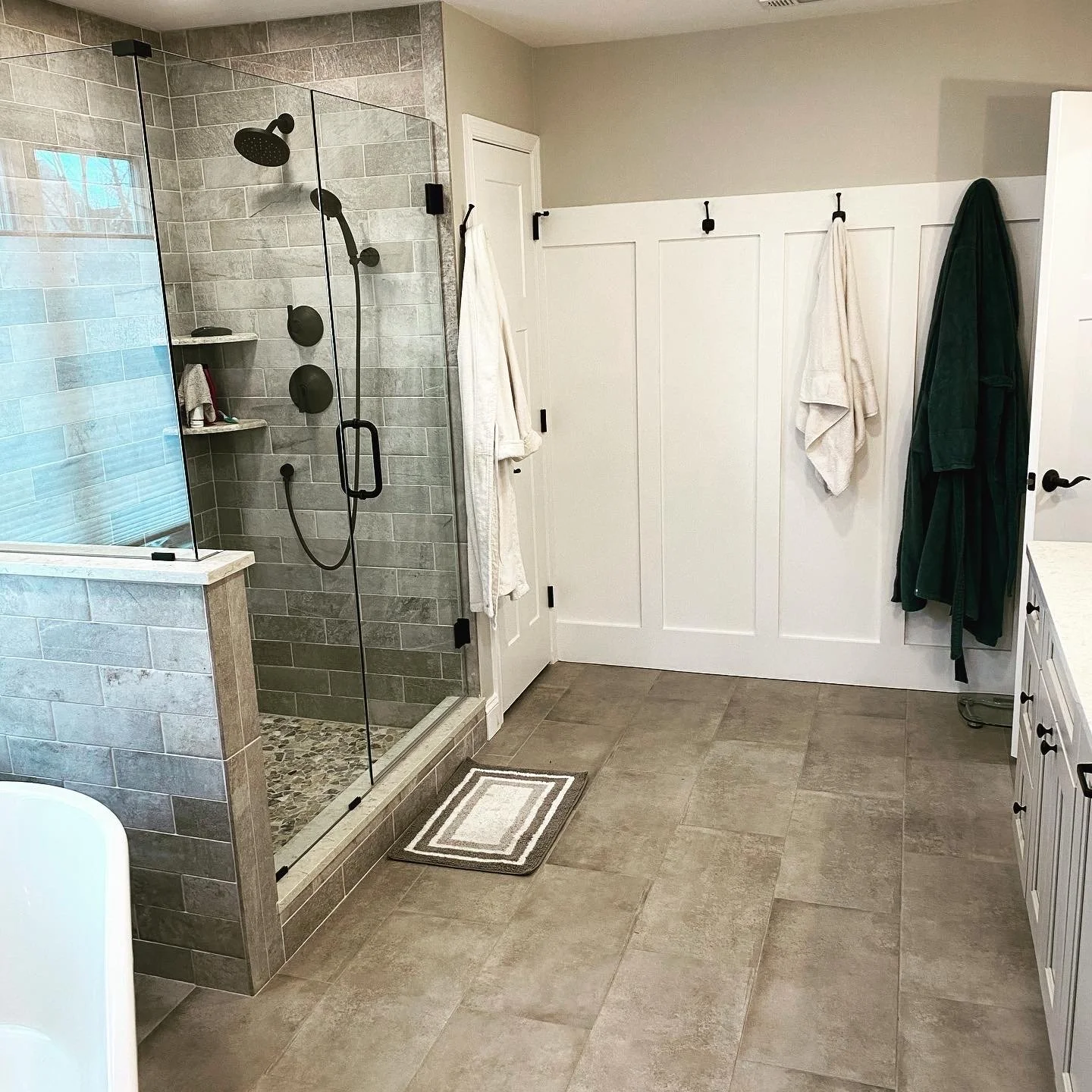 Bathroom with glass-enclosed shower, white wainscoting on the wall, and a beige tiled floor. There are towels hanging on hooks and a green bathrobe hanging on the wall.