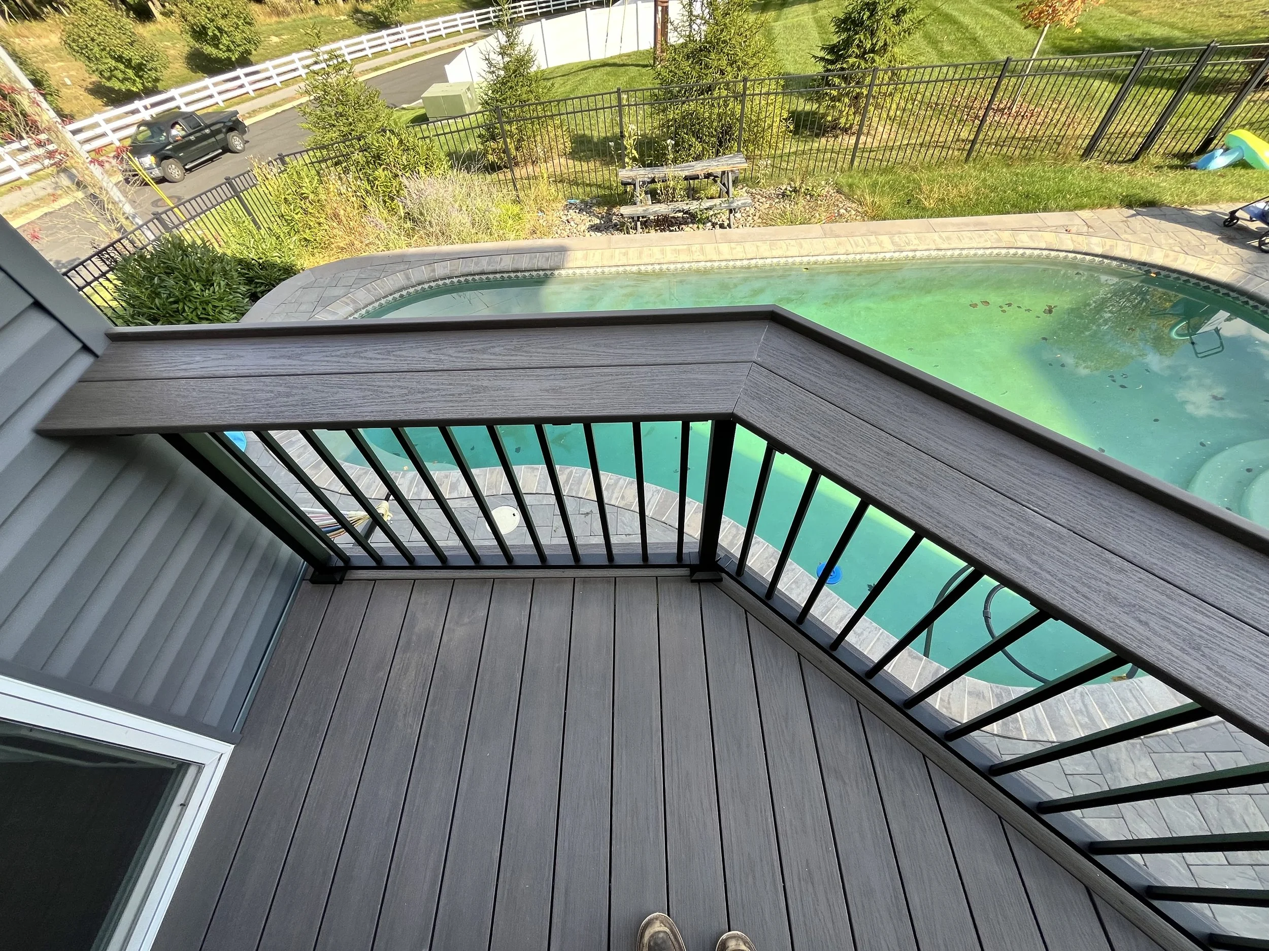 View of a backyard pool area from a balcony with a wooden railing, showing the greenish water in the pool, a nearby patio with tiles, a grassy yard with trees, a white fence, a parked black pickup truck, and a winding asphalt road.