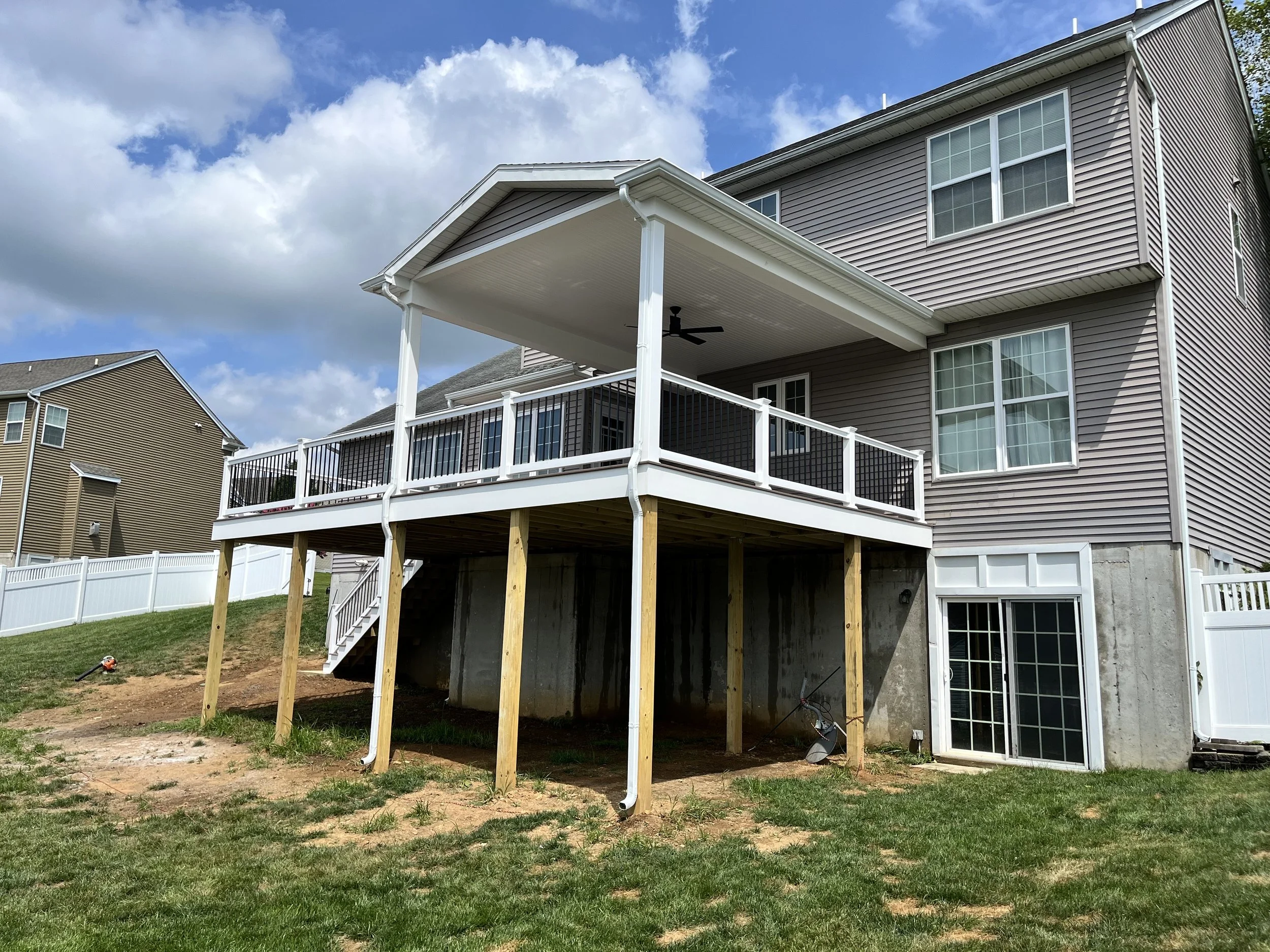 Backyard with raised deck under construction, supported by wooden posts, with a house featuring a screened-in porch, windows, and siding, during daytime with partly cloudy sky.