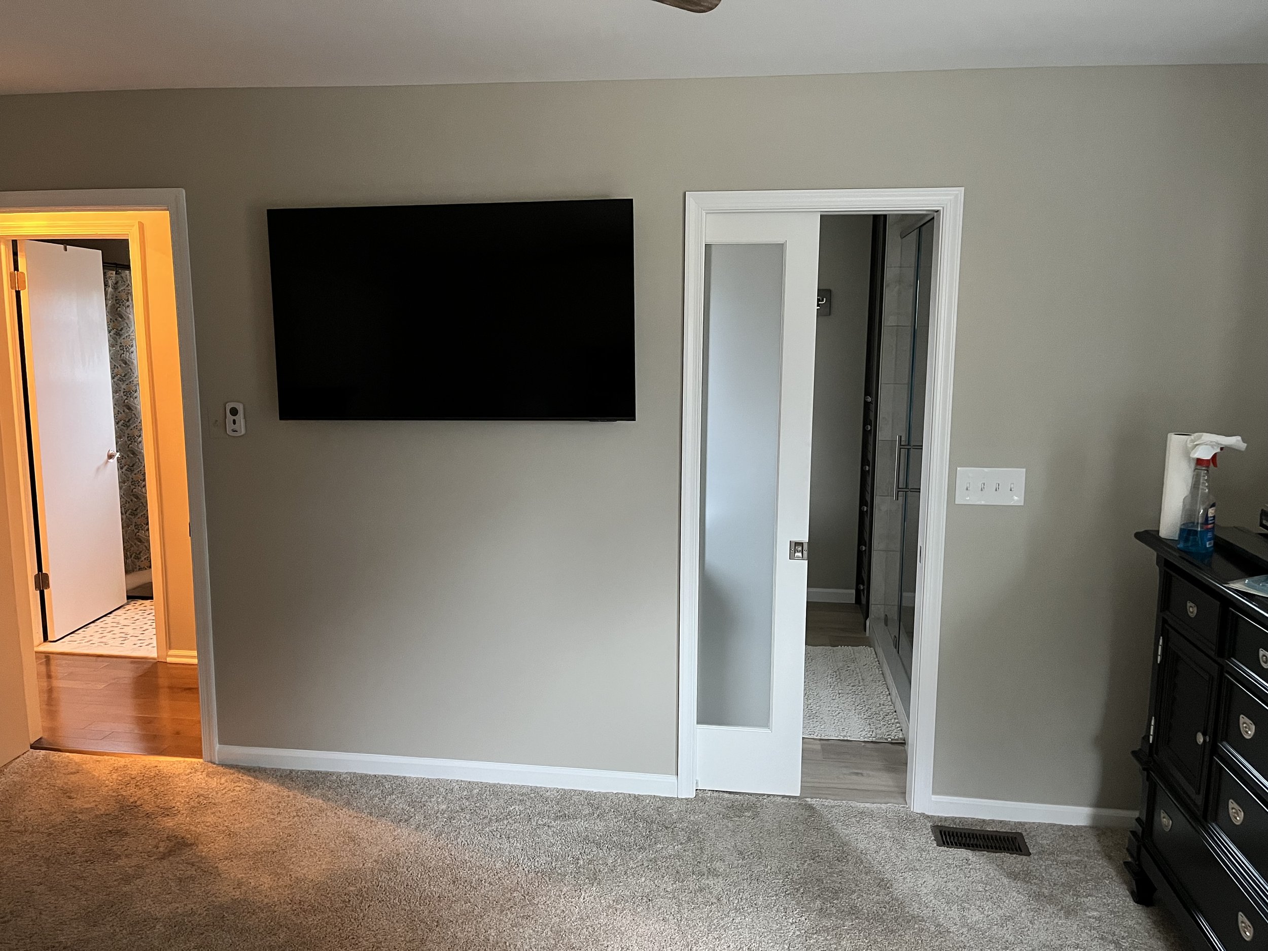 Interior view of a room with a wall mounted TV, a doorway with white trim leading to a bathroom, and a black dresser on the right side, with some cleaning supplies on top.