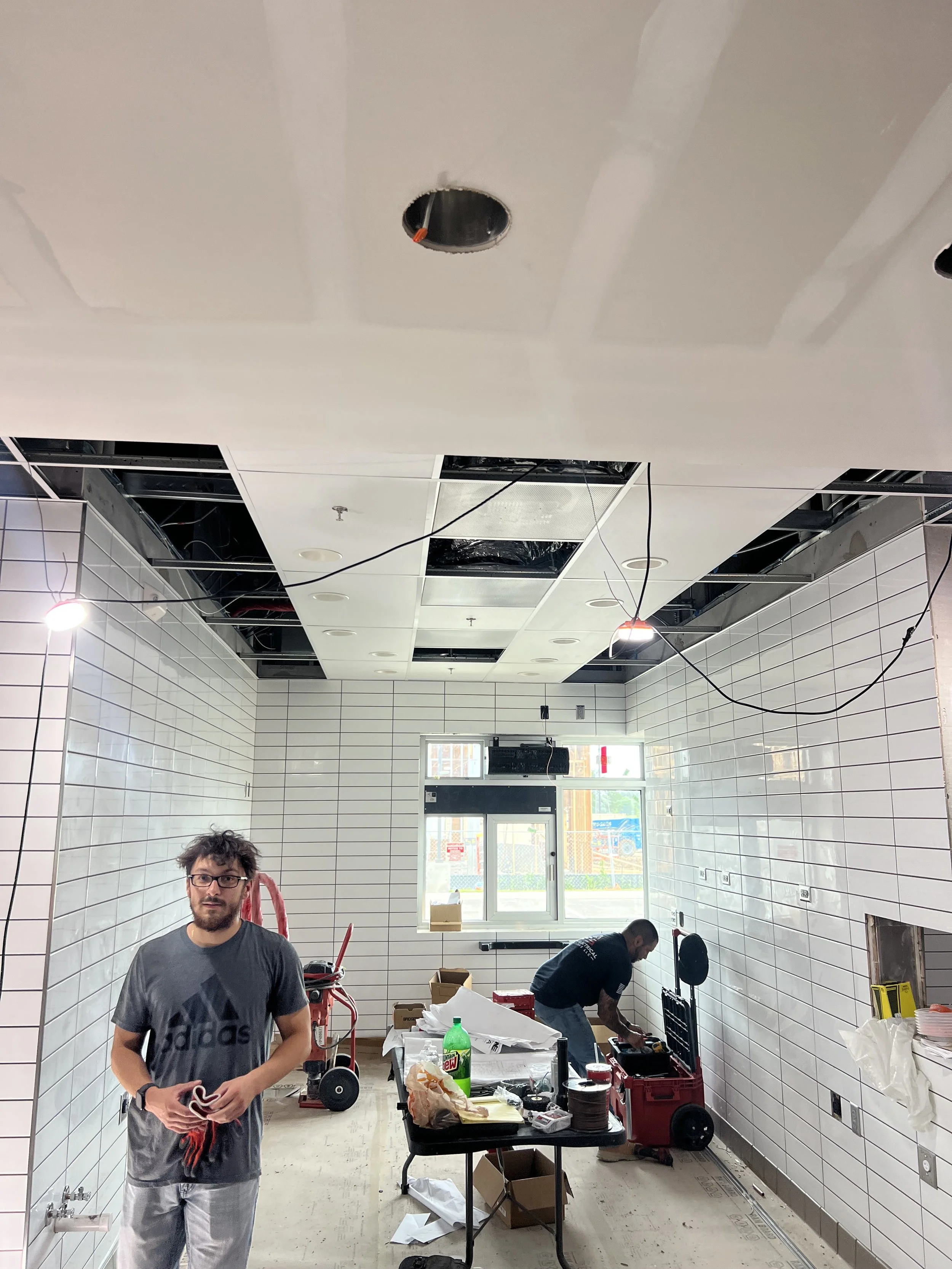 Two men are working in a room with white tiled walls. One man with glasses and a beard, wearing a gray Adidas t-shirt, is standing near the camera and holding some objects. Another man is crouching down, working on something near the wall. The room appears to be under construction or renovation, with exposed ceiling panels, wires, and tools scattered on the table and floor.