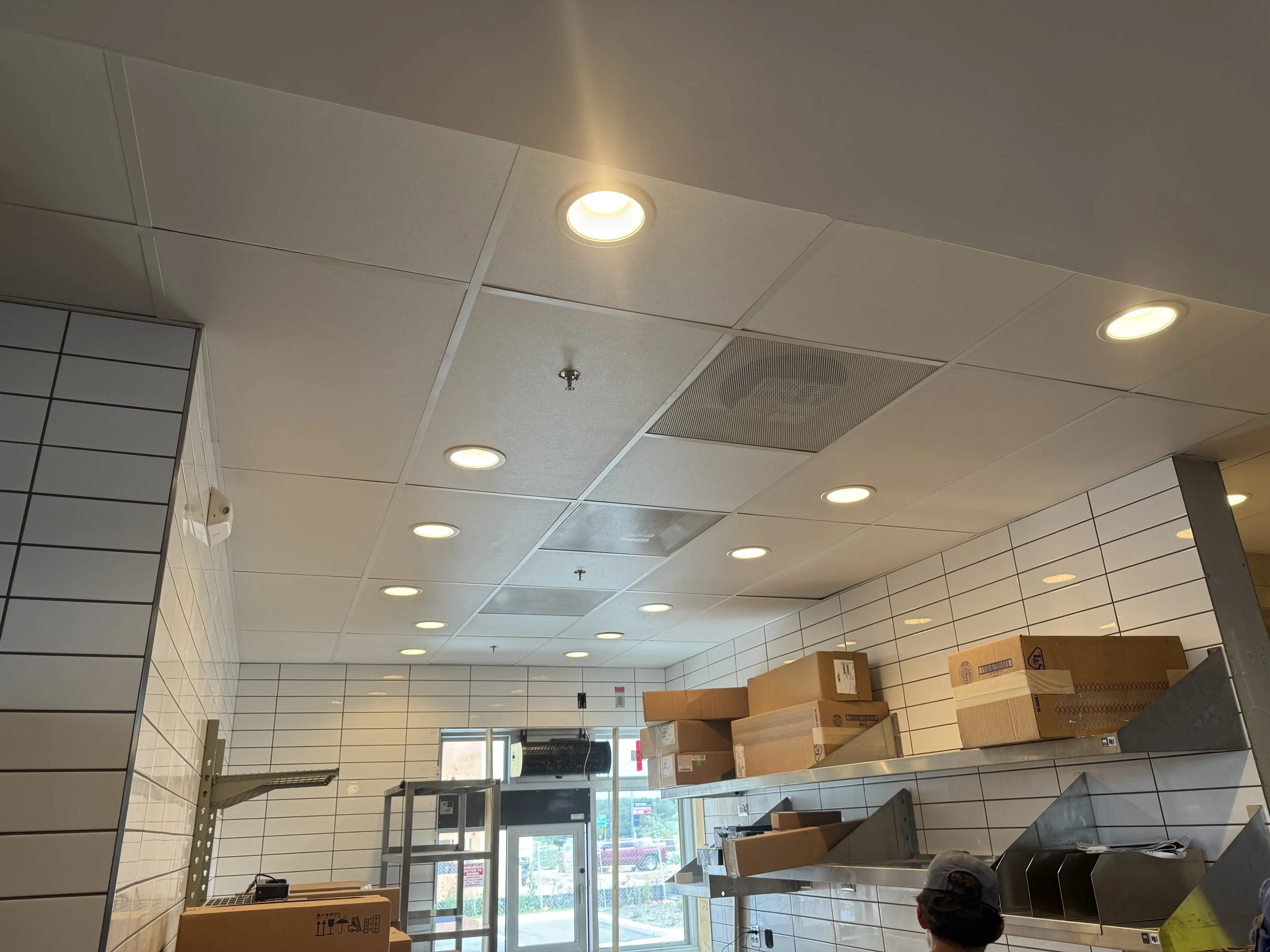 Ceiling with multiple recessed lights in a commercial or retail space, partially visible boxes on tables, and a door/window in the background.