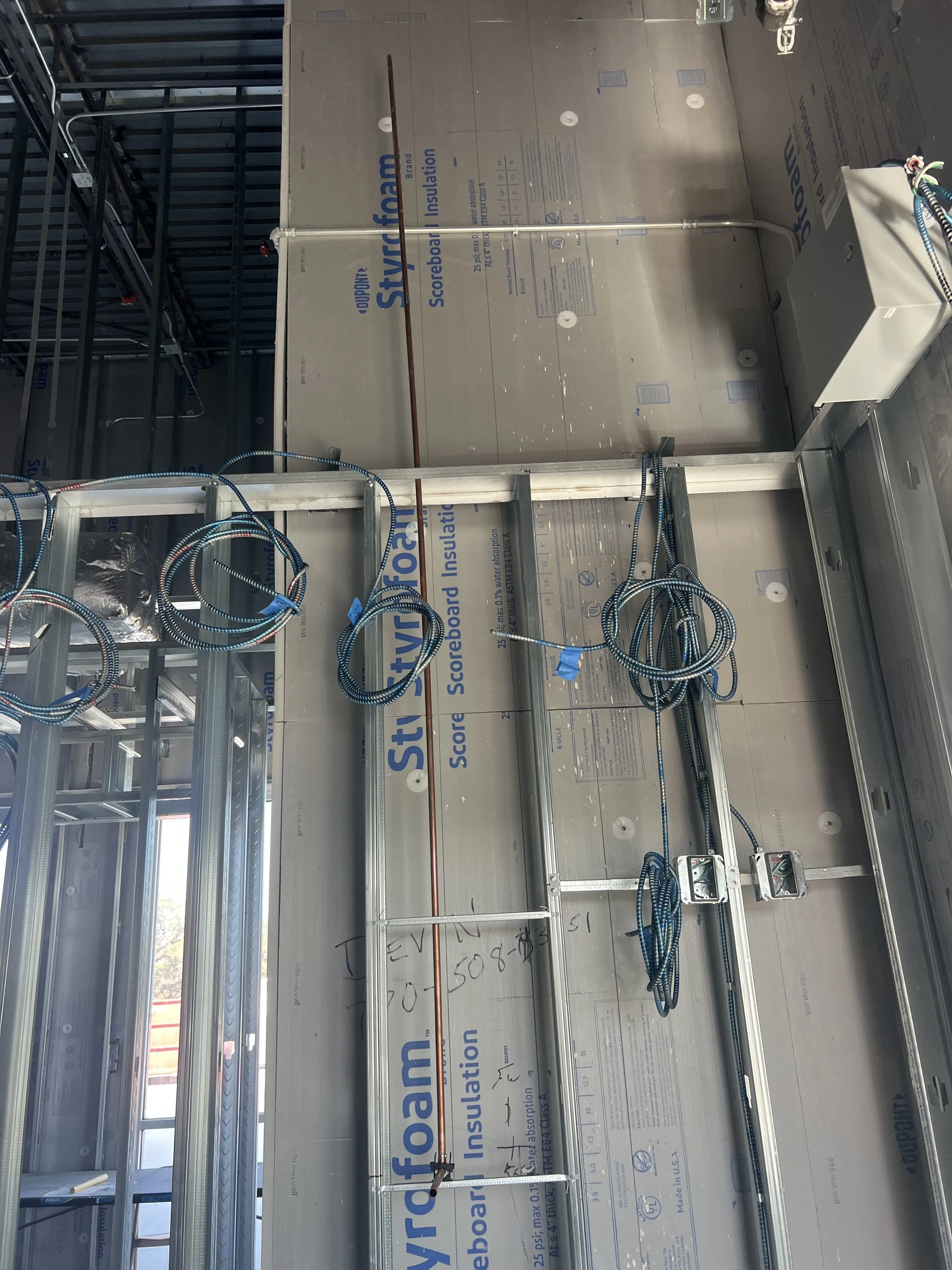 Interior of a building under construction showing metal framing with electrical wiring, electrical boxes, and drywall panels with insulation markings.