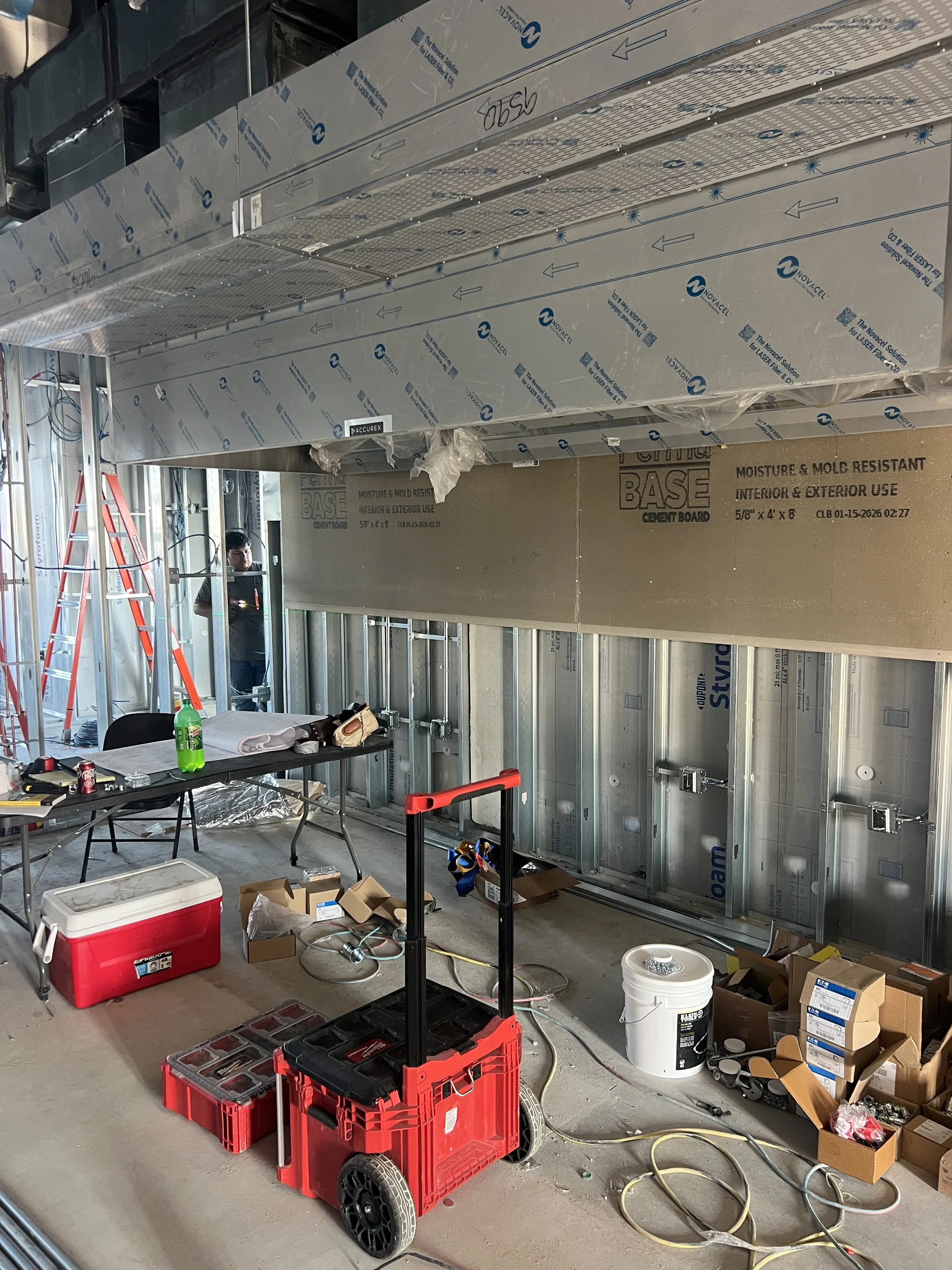 Construction site showing metal wall framing, insulation panels, and ceiling tiles with tools, boxes, coolers, and construction materials scattered on the floor.