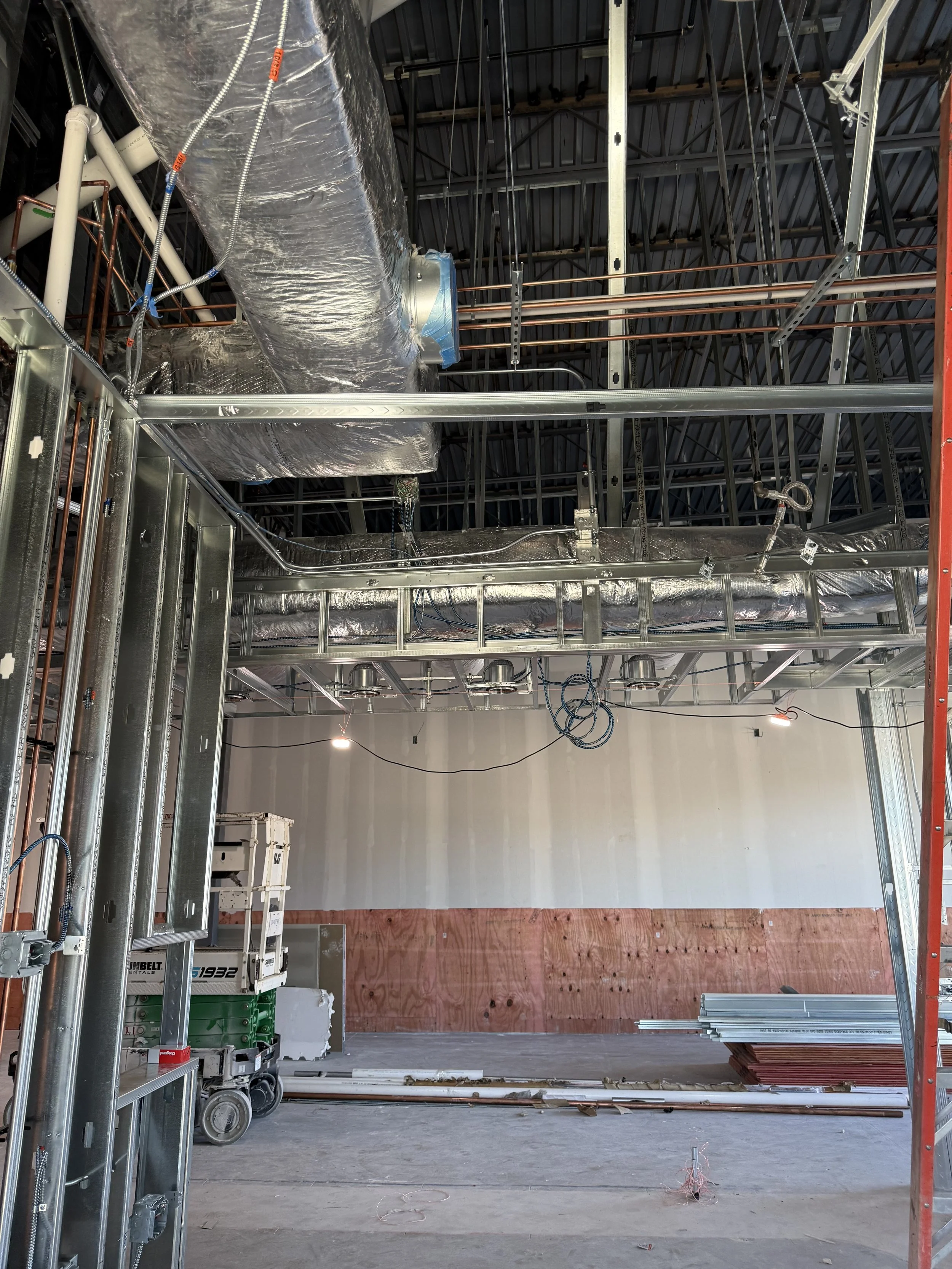 Interior of a building under construction, showing exposed metal framing, ductwork, pipes, and partially finished walls.