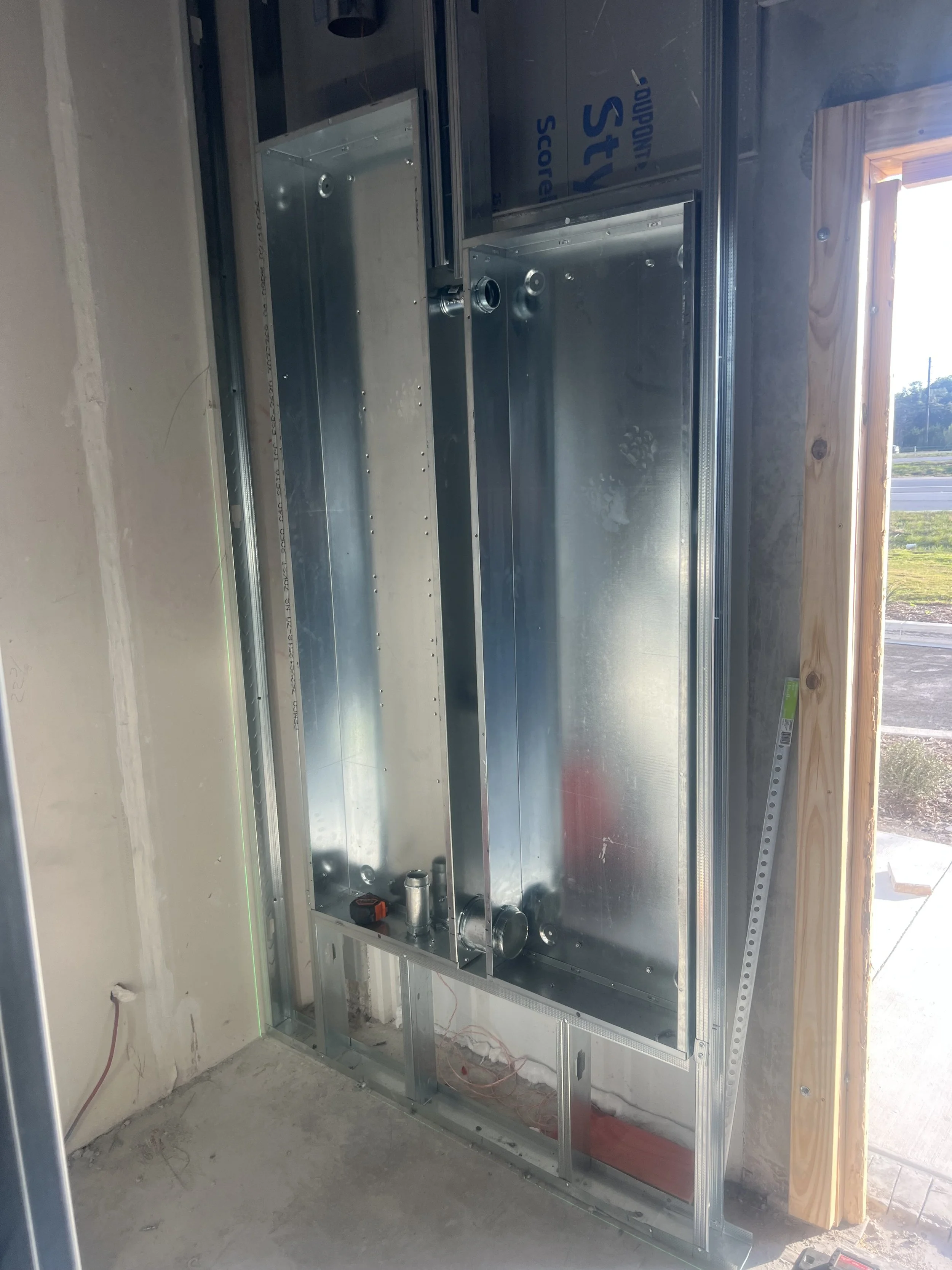 Metal framing and plumbing inside a wall under construction, with an open doorway and a window to the outside.