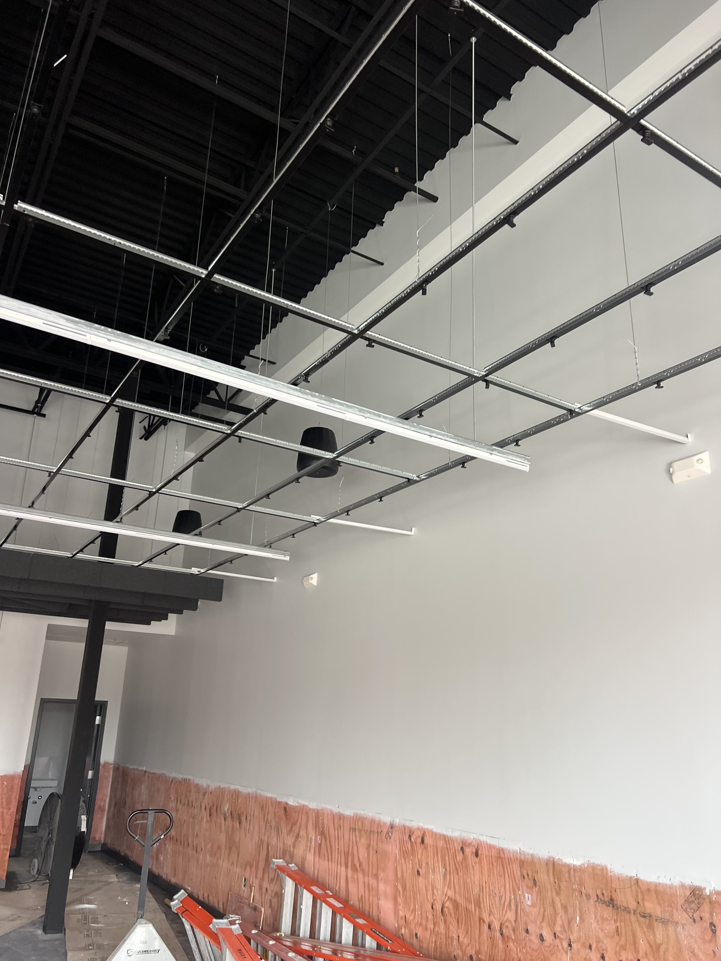 Construction or renovation site inside a building with a high ceiling, with hanging metal tracks and lighting fixtures, a ladder, a partially finished wall, and construction tools.