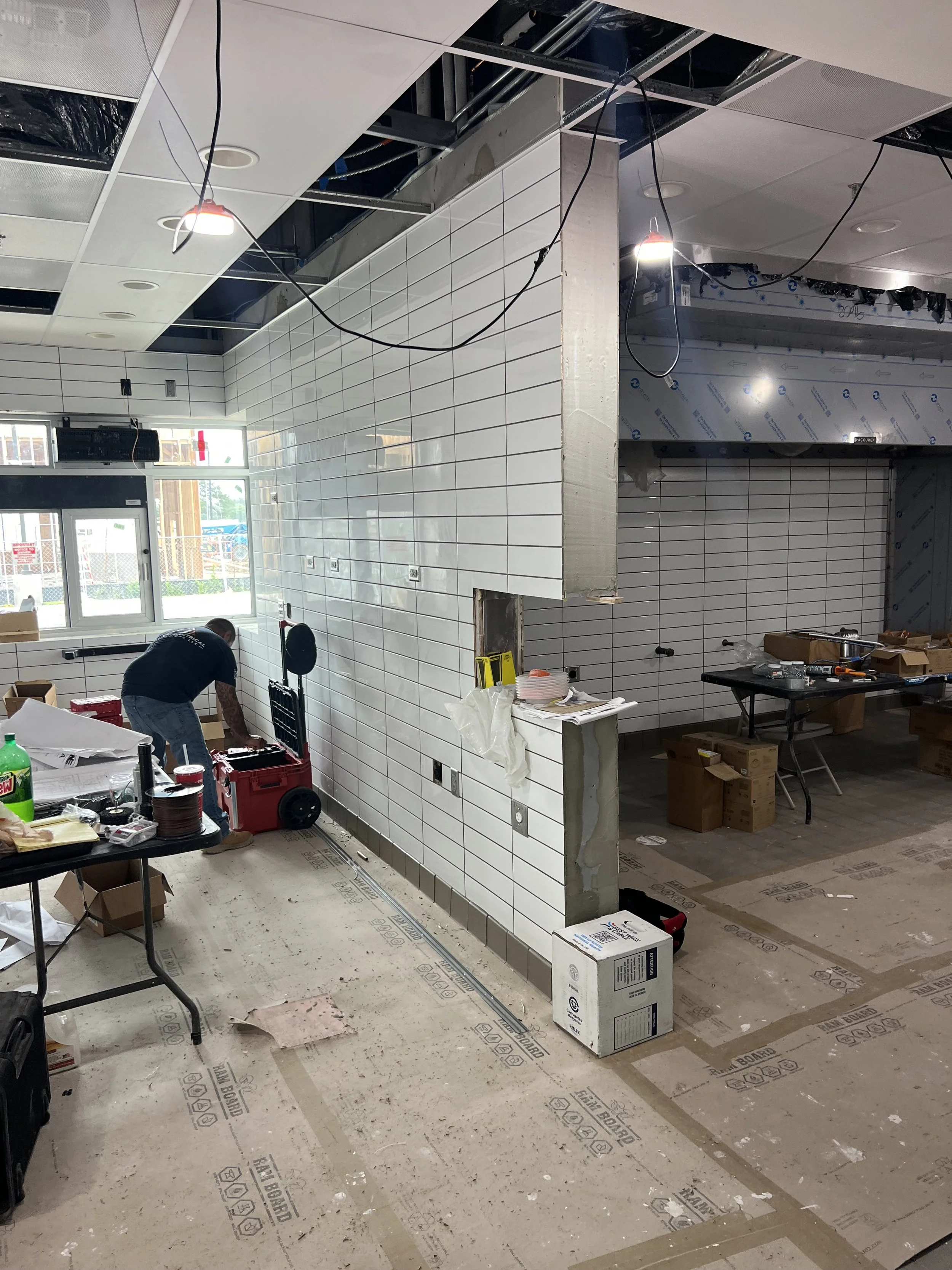 Interior of a commercial space under construction with partially installed white tiles on the wall, construction tools and supplies on tables and floor, and workers installing or renovating the space.