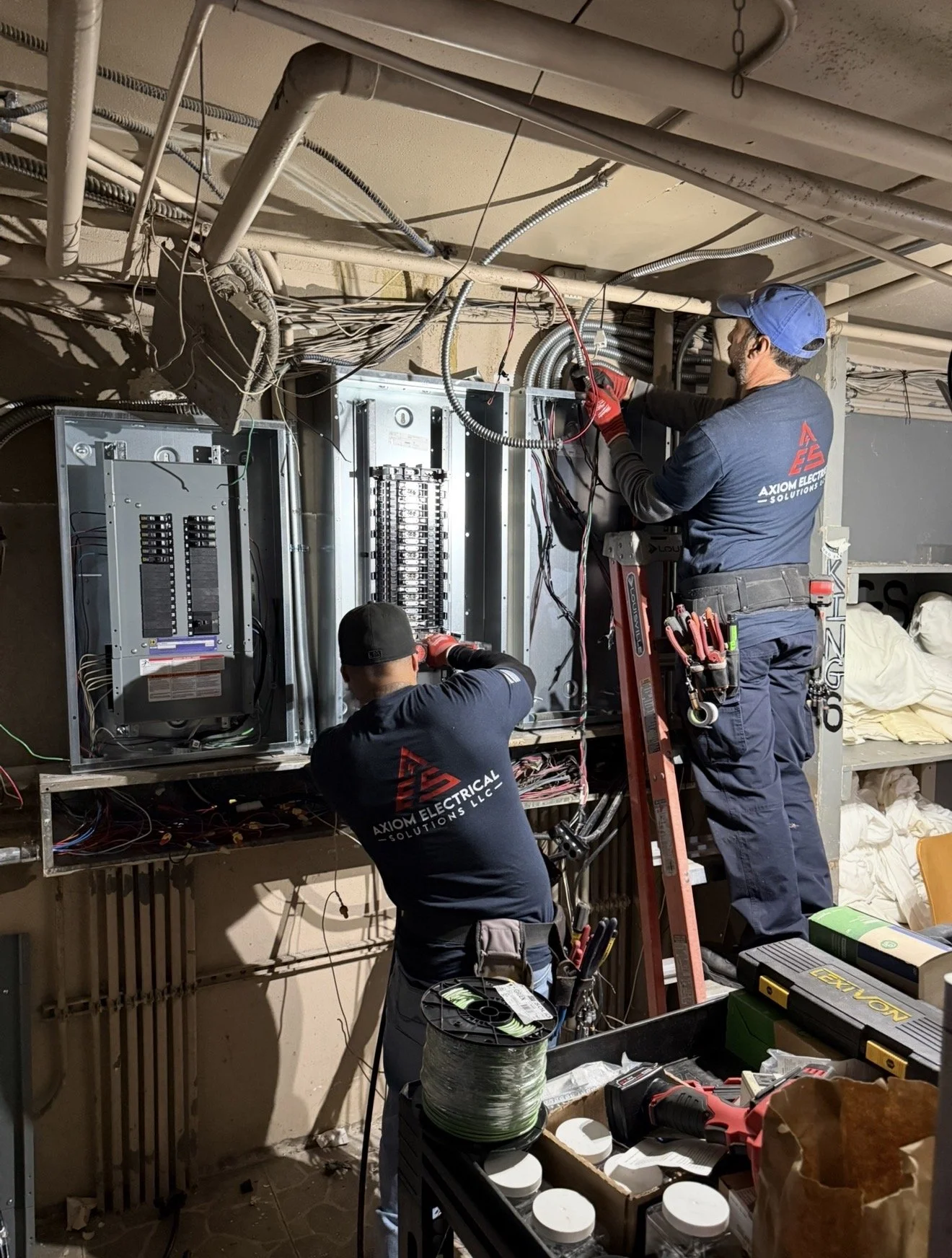 Two electricians working on electrical panels, with tools and wiring around them, in an indoor setting.