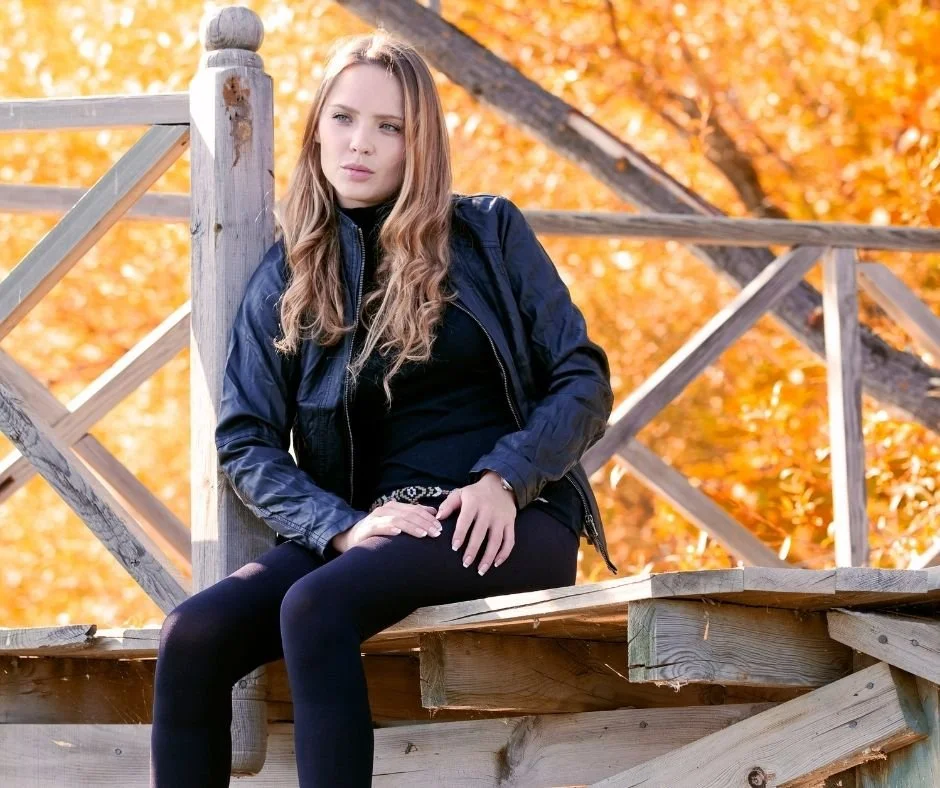 Woman in a dark jacket sitting on a wooden railing, gazing off to the side, with vivid orange autumn foliage in the background