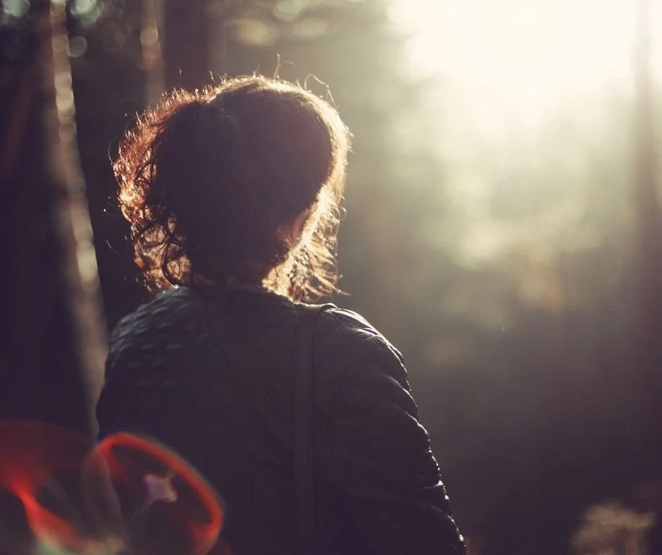Woman with curly hair walking alone through sunlit trees, seen from behind.