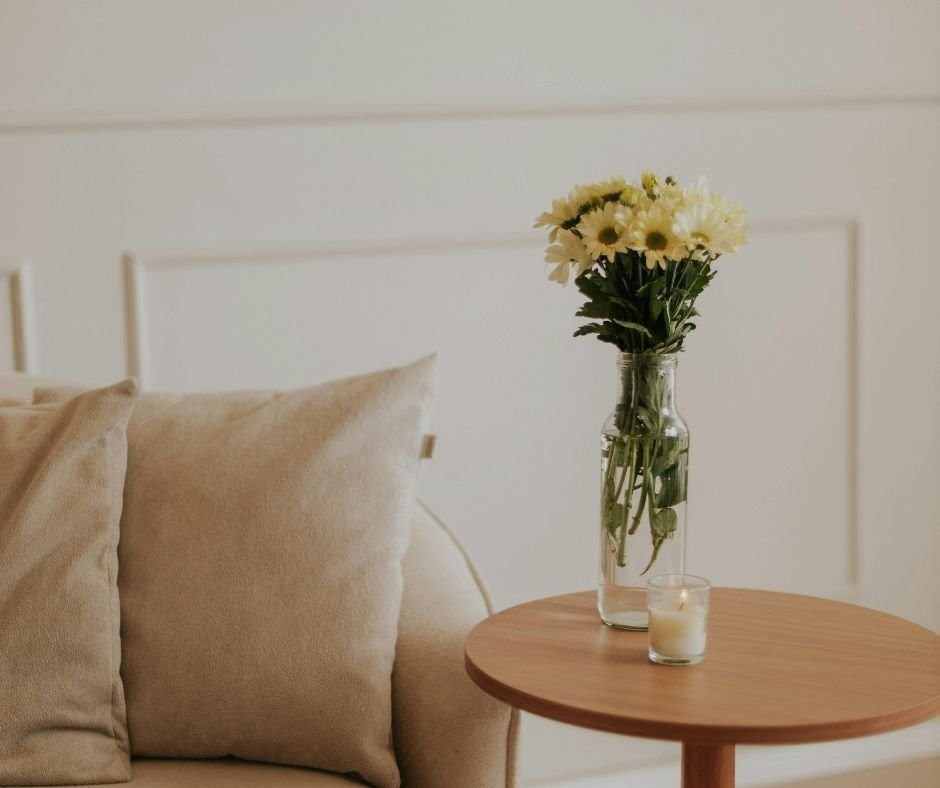 A jar of white daisies and a lit candle on a small wooden side table, beige throw pillows on a sofa in the background