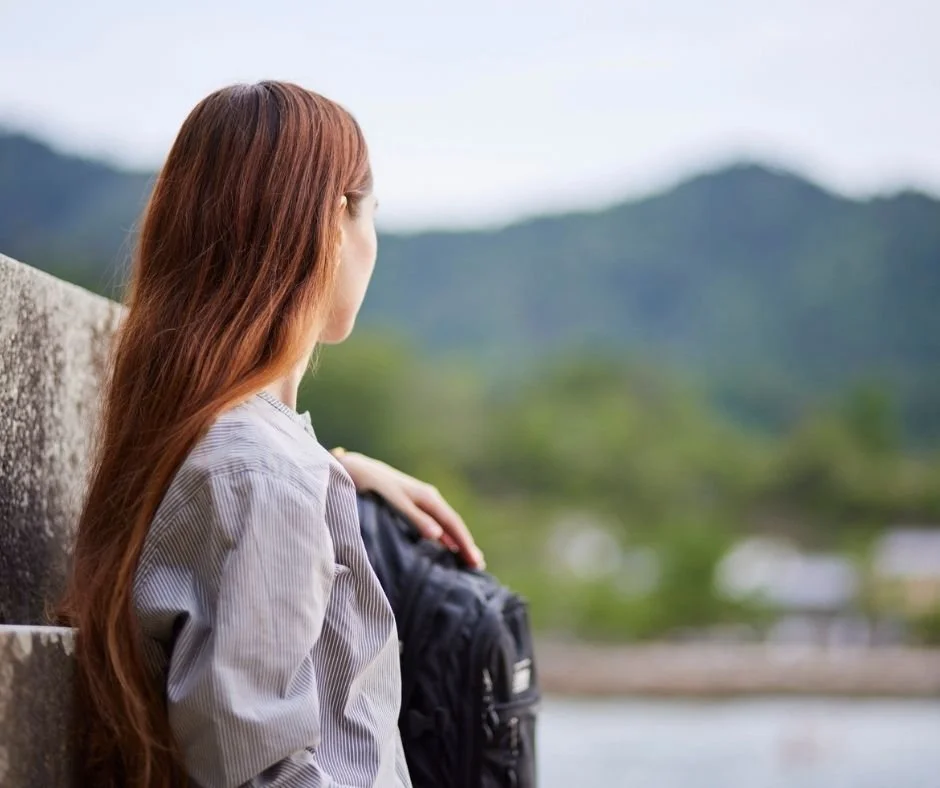 Woman with a backpack leaning against a stone wall, looking out at hills across the water