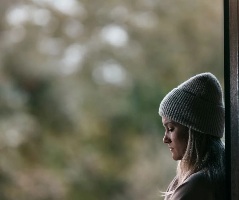 Woman in a gray knit beanie looking down pensively in a doorway, soft blurred greenery in the background