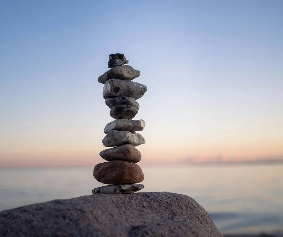 Stack of balanced stones on a rock at the shoreline, with the ocean and sunset sky in the background.