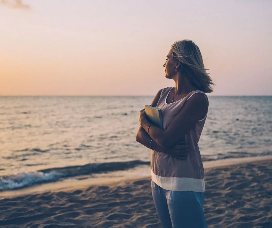 Woman pausing by the ocean at sunset, holding a journal in quiet reflection, what to expect in therapy at Bountiful Health