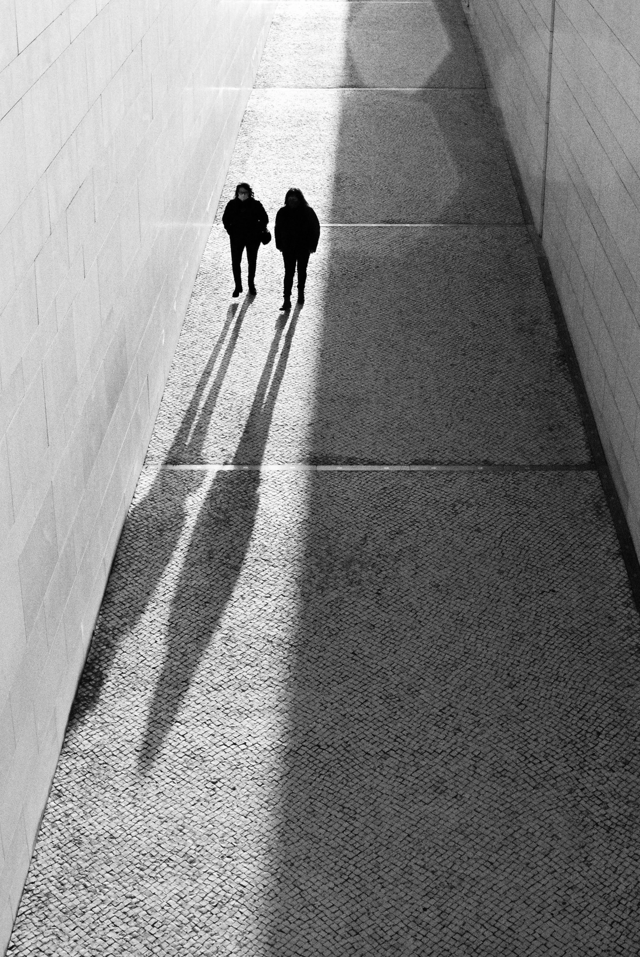 Two figures walking side by side through a sunlit passage, casting long parallel shadows.