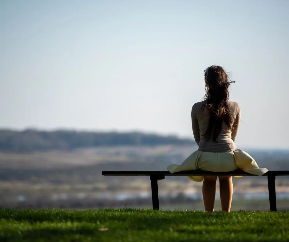 Woman seen from behind, sitting alone on a bench overlooking rolling green hills and open sky