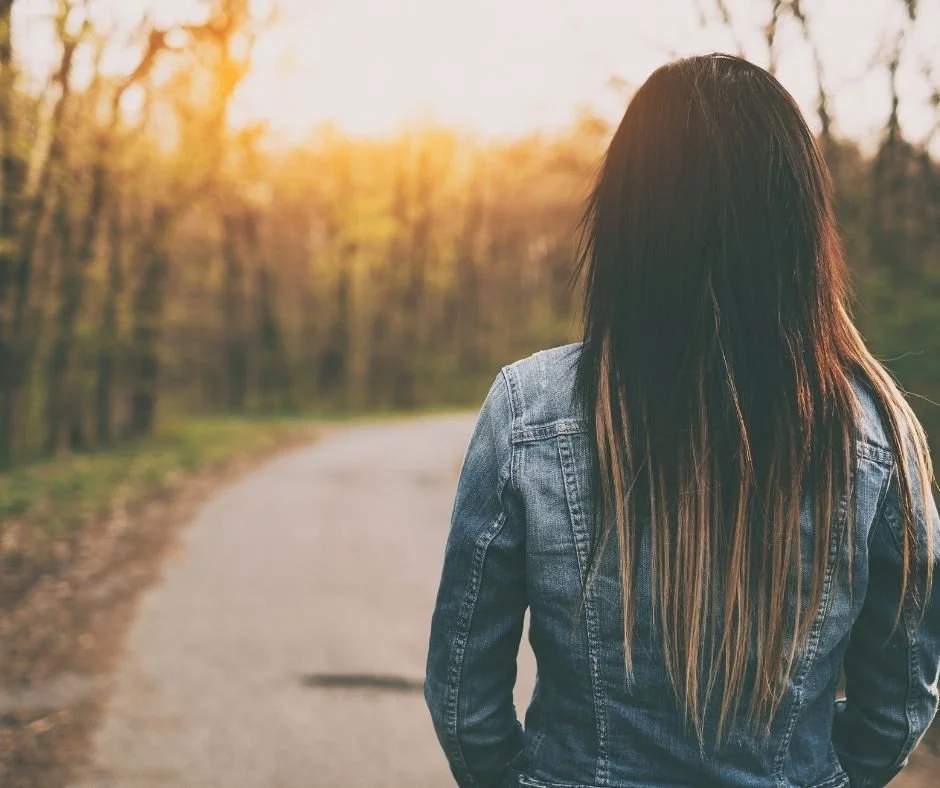 Woman in a denim jacket walking away down a sunlit path through trees, therapy in San Ramon, CA