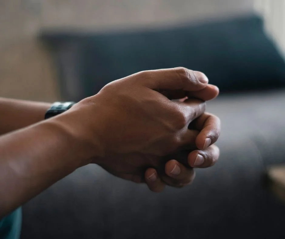 Close-up of a person's clasped hands, seated in a dimly lit room with a couch and cushion in the background