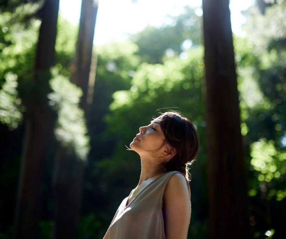 Woman standing among tall redwood trees with eyes closed, breathing in dappled sunlight, therapy in San Ramon, CA