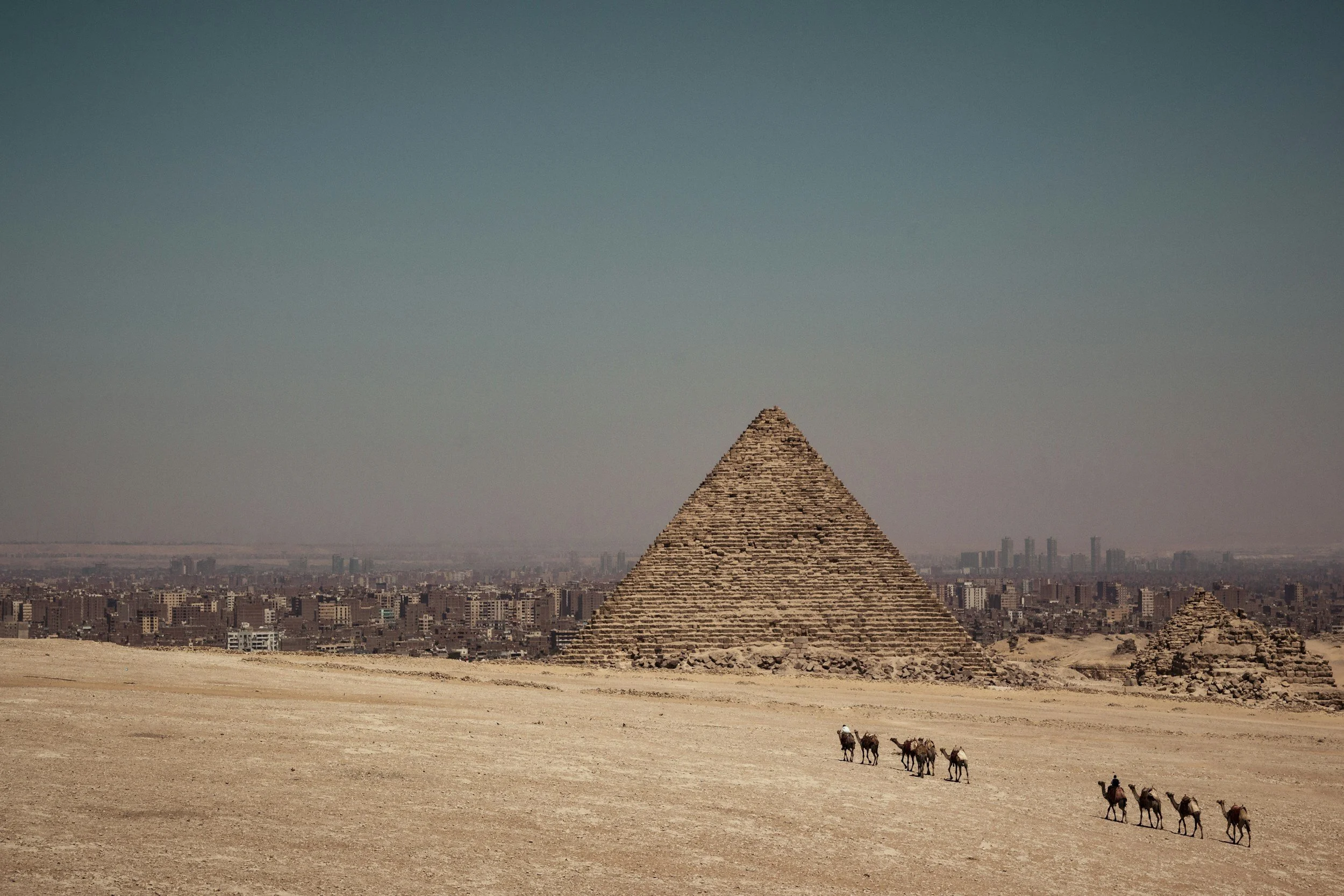 The Great Pyramid of Giza in Egypt with a group of camels and a traveler in the foreground, city skyline in the background under clear sky.