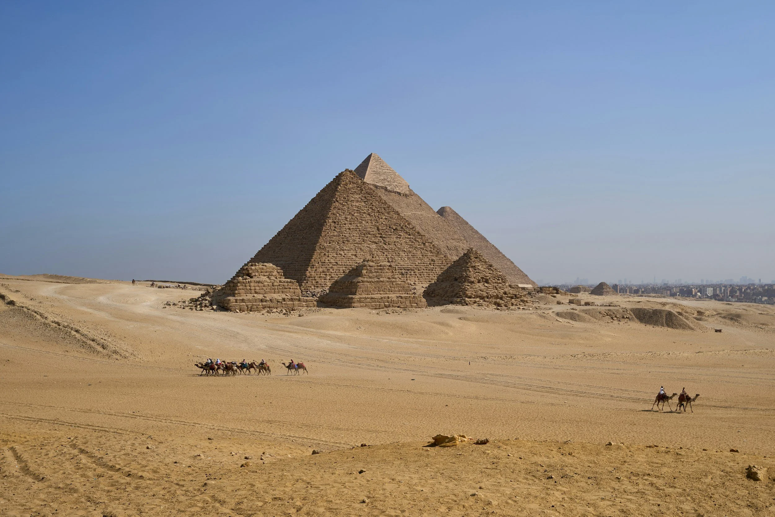 The ancient pyramids of Egypt in a desert landscape with a clear blue sky, and a group of camels with people riding them in the foreground.