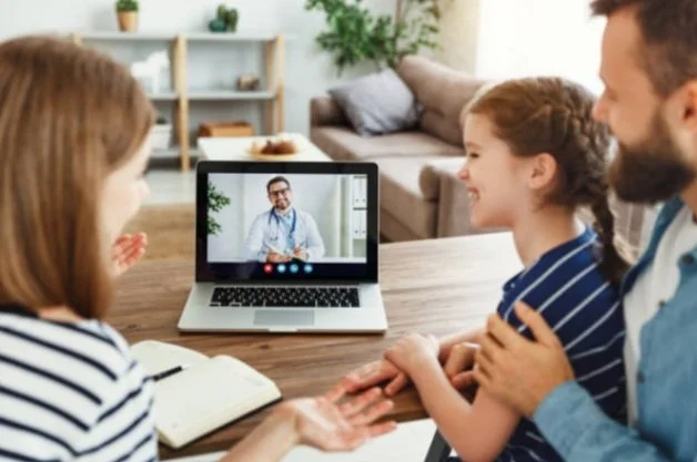A family participating in a telehealth appointment with a doctor on a laptop in a living room.
