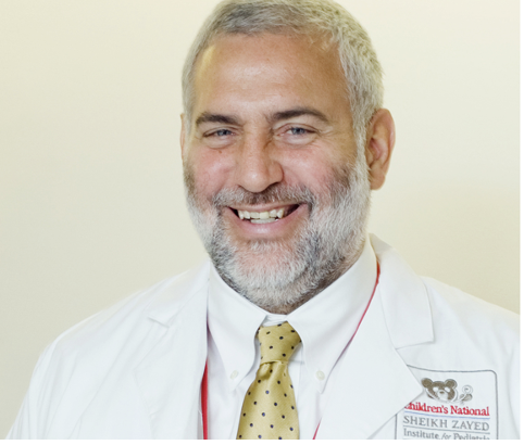 A smiling male doctor with gray hair and a beard, wearing a white coat and a yellow tie, in a professional setting.