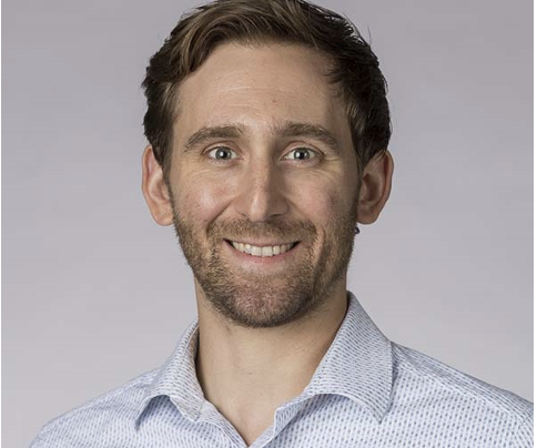 A smiling man with short brown hair and a beard, wearing a light blue dress shirt, against a plain gray background.