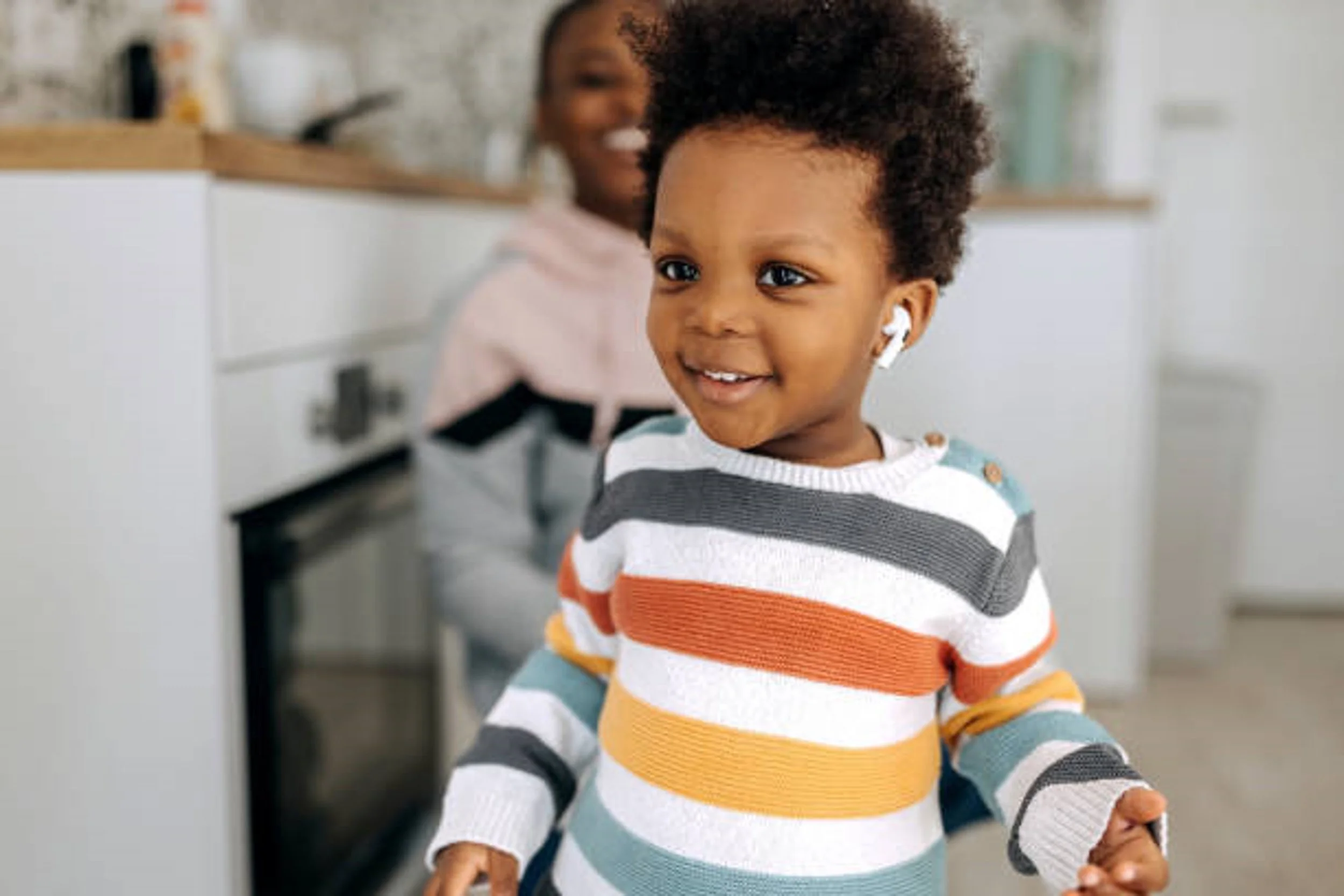 A smiling young girl with curly hair wearing a colorful striped sweater, with a second girl in the background, both in a modern kitchen.